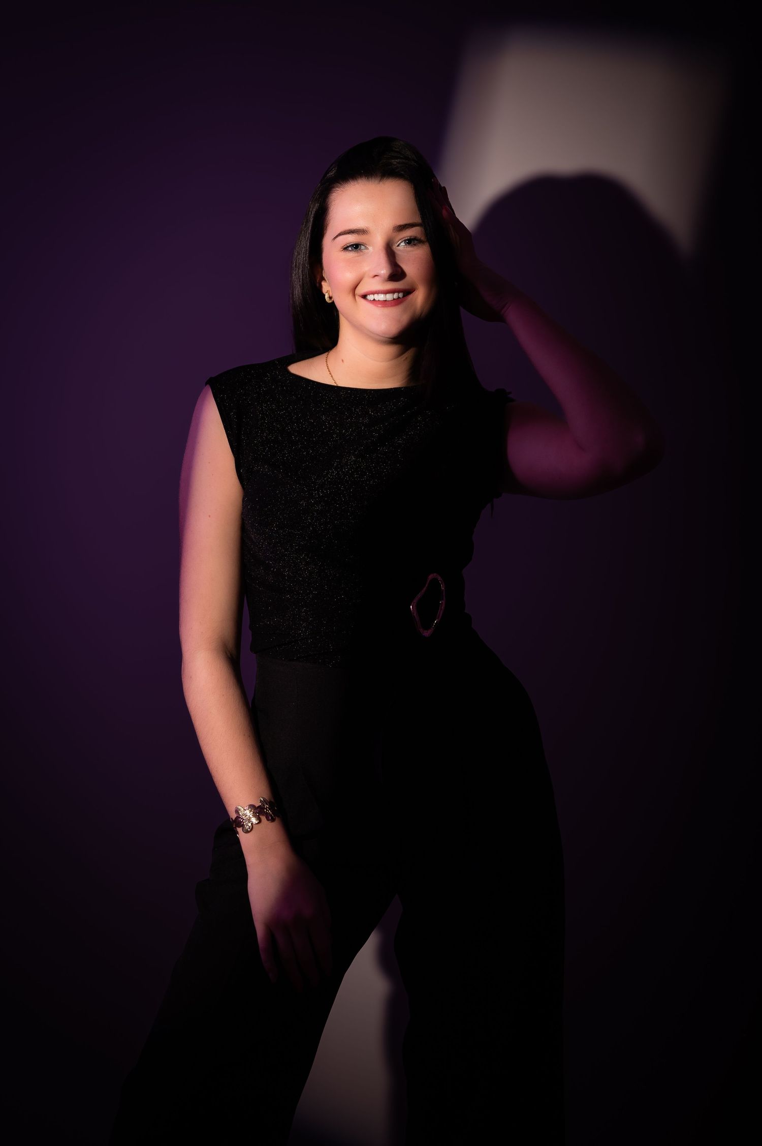 Young woman in black sleeveless dress smiling confidently against a purple studio backdrop.