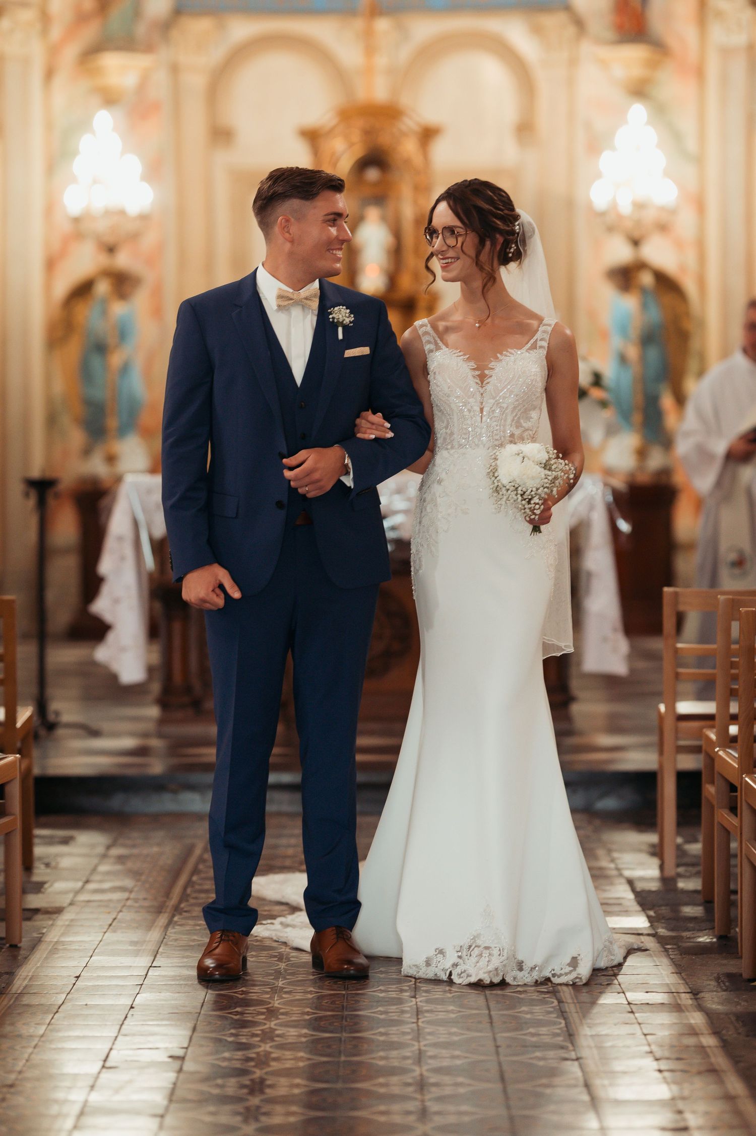 Bride and groom walking down church aisle, she in lace mermaid gown, he in navy suit with bow tie.
