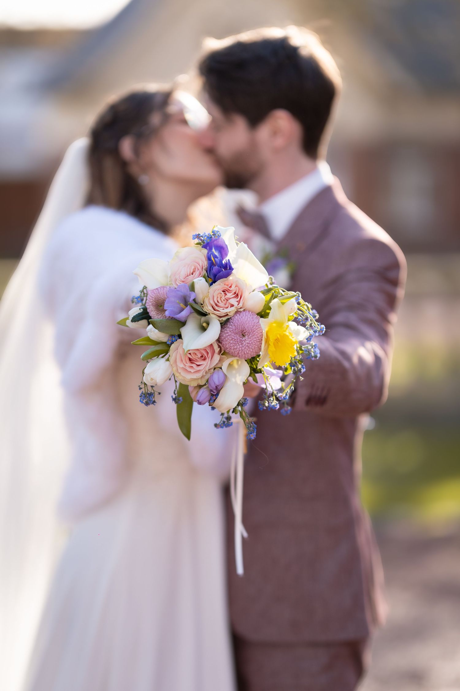 Bride and groom kissing outdoors with a vibrant spring bouquet of roses and wildflowers in focus.