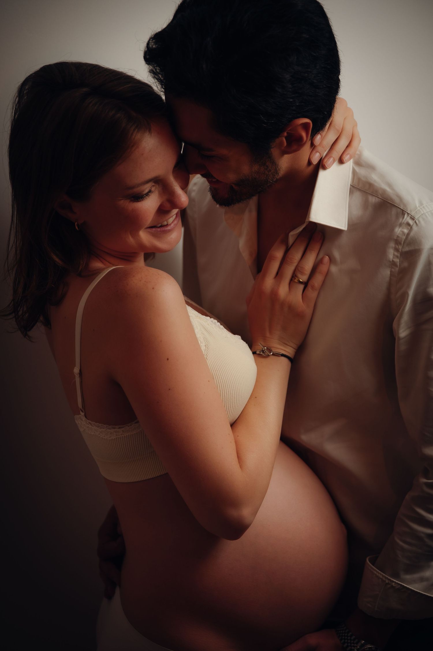 Couple sharing tender maternity moment, pregnant woman in cream bra embraced by partner, forehead touch pose.