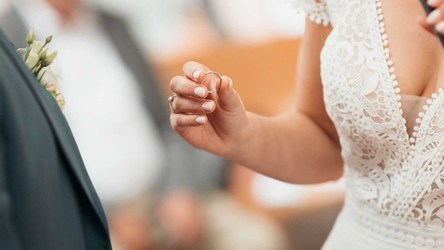 Bride and groom holding hands during wedding ceremony, bride wearing white lace dress with floral boutonniere visible.