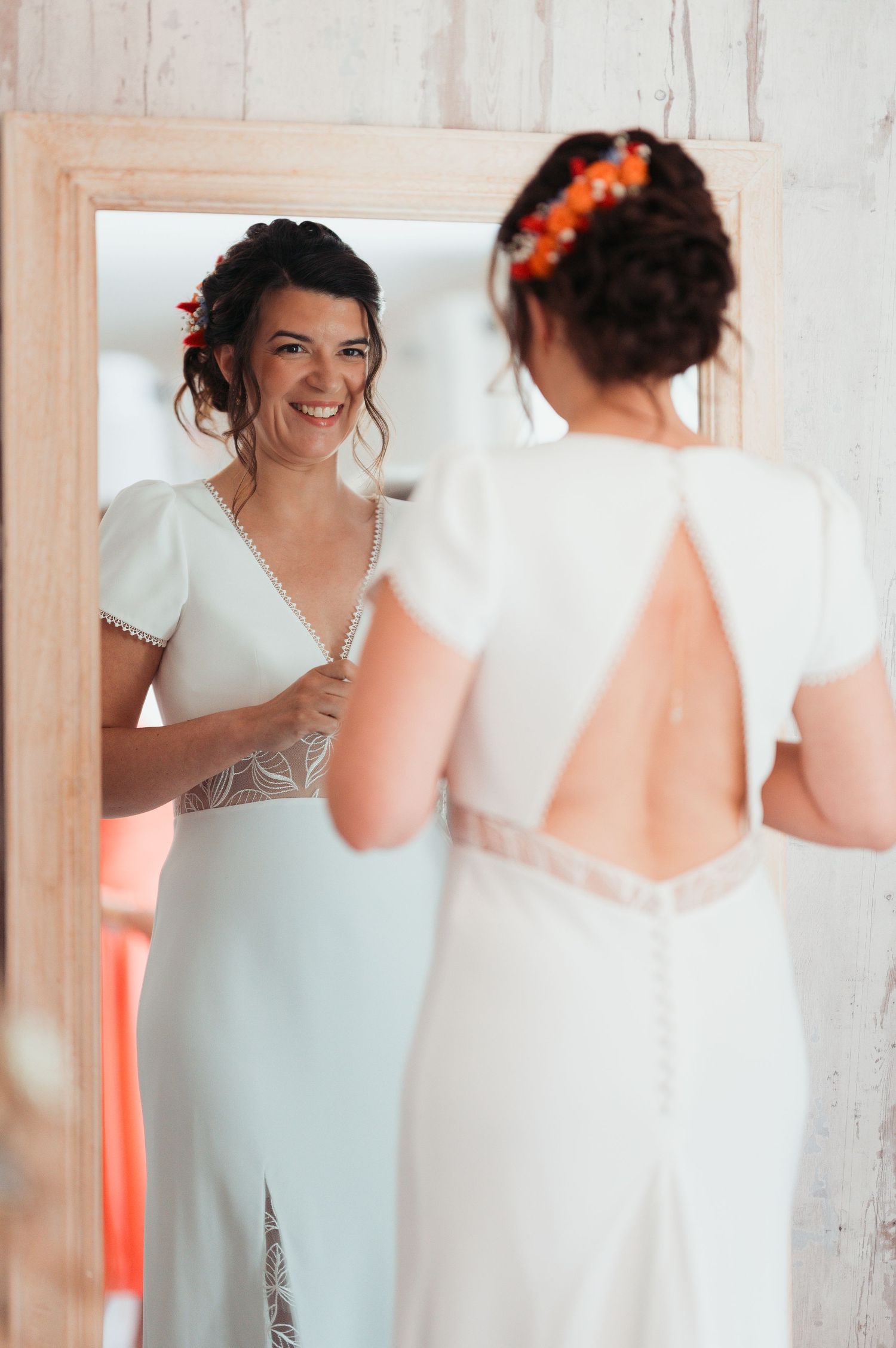 Smiling bride in white open-back wedding dress with orange floral hair accessory admiring herself in a wooden mirror.