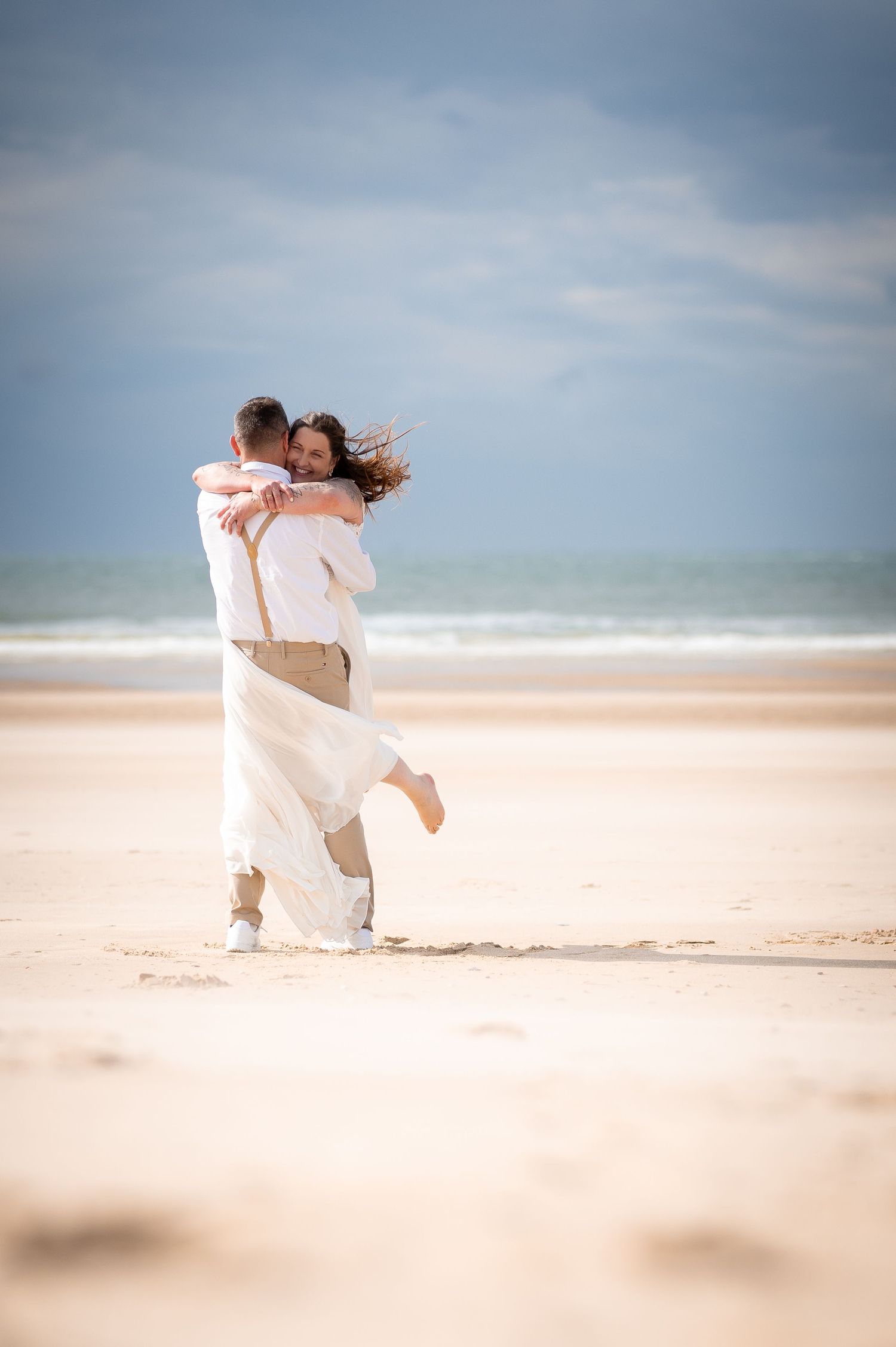 Couple embracing on a sandy beach, woman lifted in the air, wearing white outfits with ocean waves in the background.