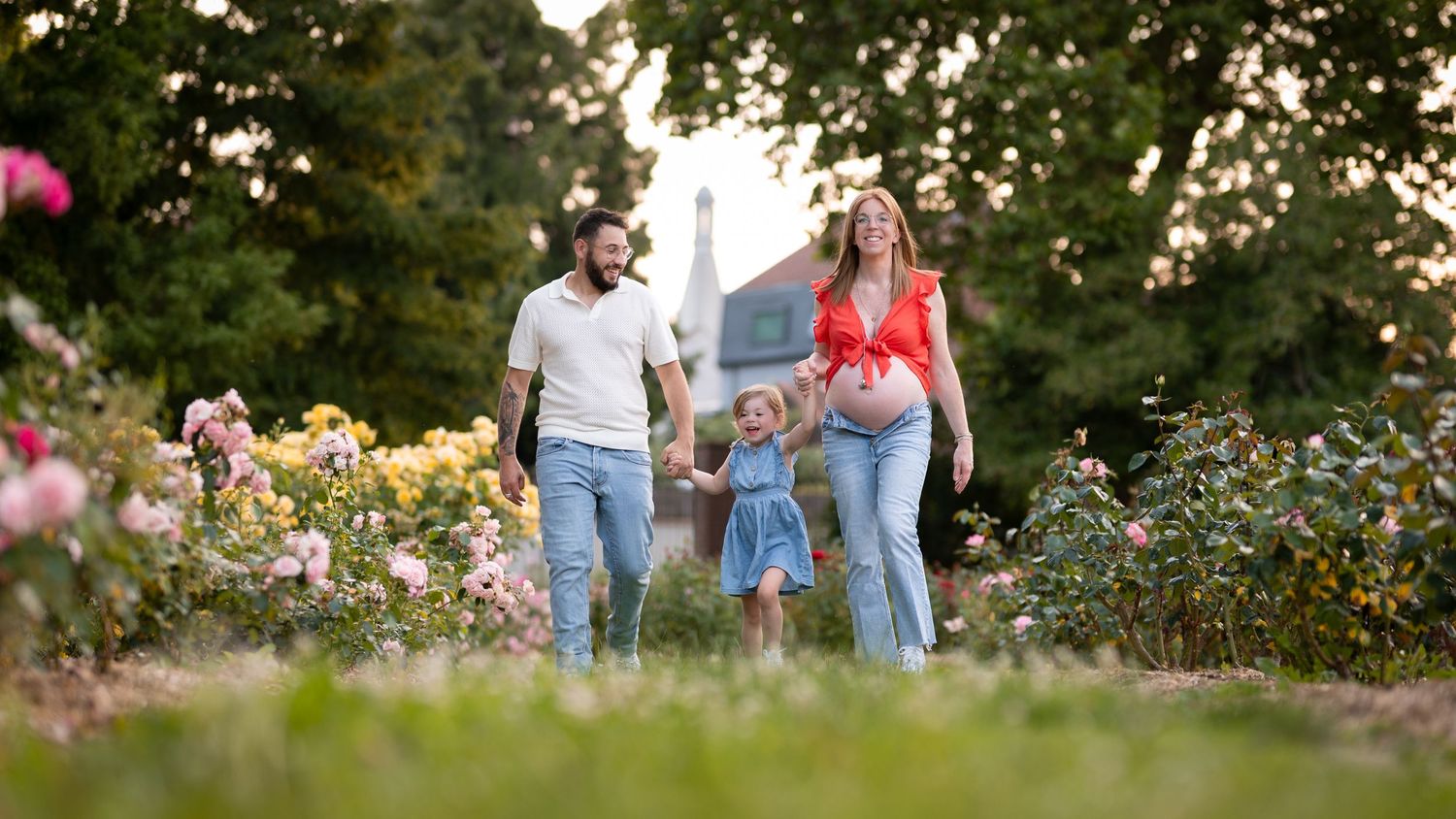 A family of three walks through a lush rose garden, with a pregnant mother in red top and jeans holding hands with child.