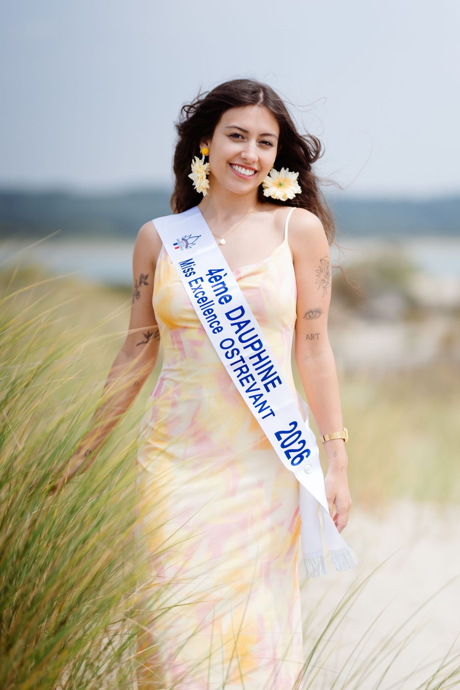 Smiling beauty queen wearing a pageant sash and floral dress walks through coastal dunes with flowers in her hair.