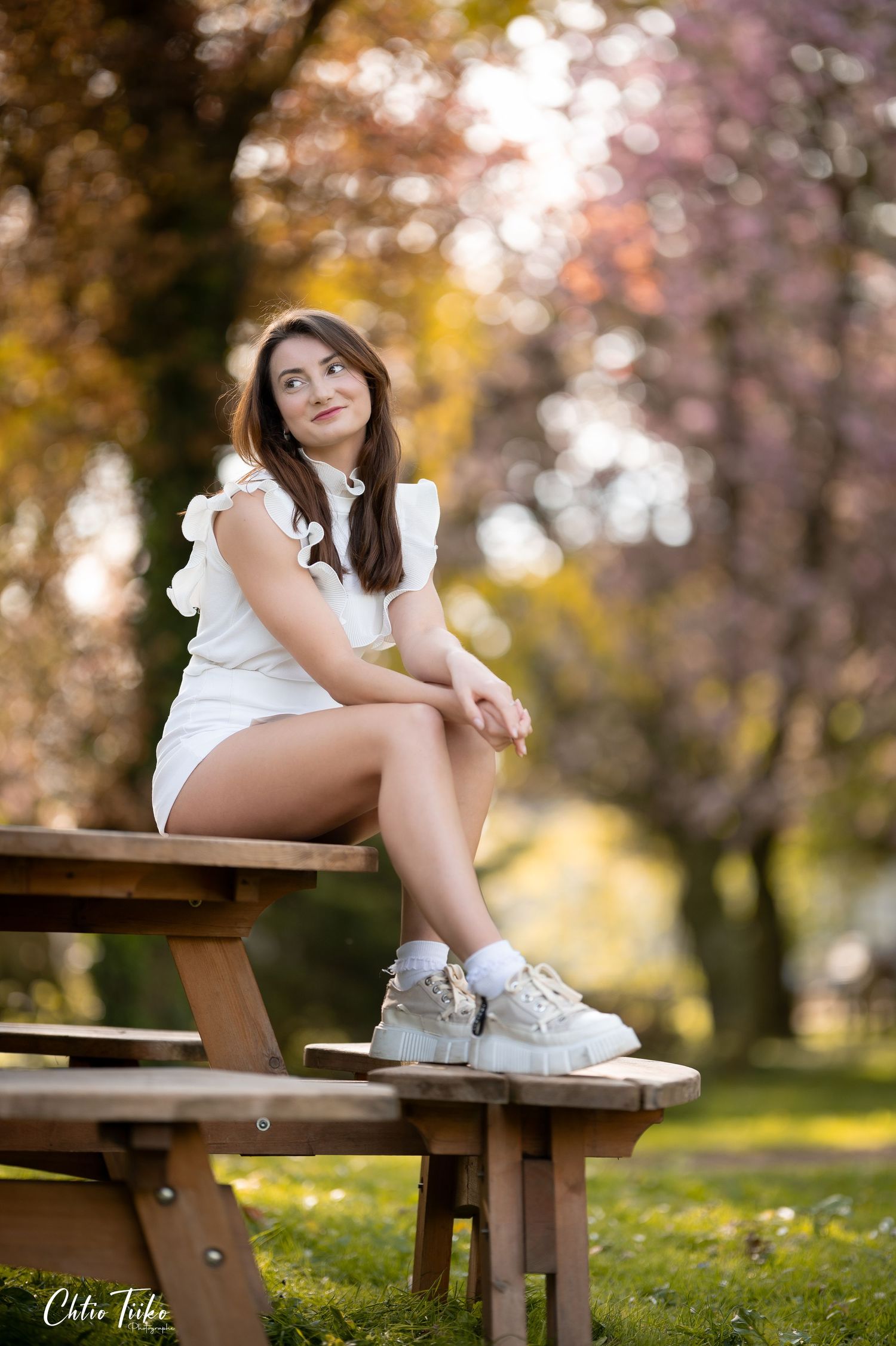 Young woman in white dress and sneakers sitting on a wooden picnic table in a blooming park setting.