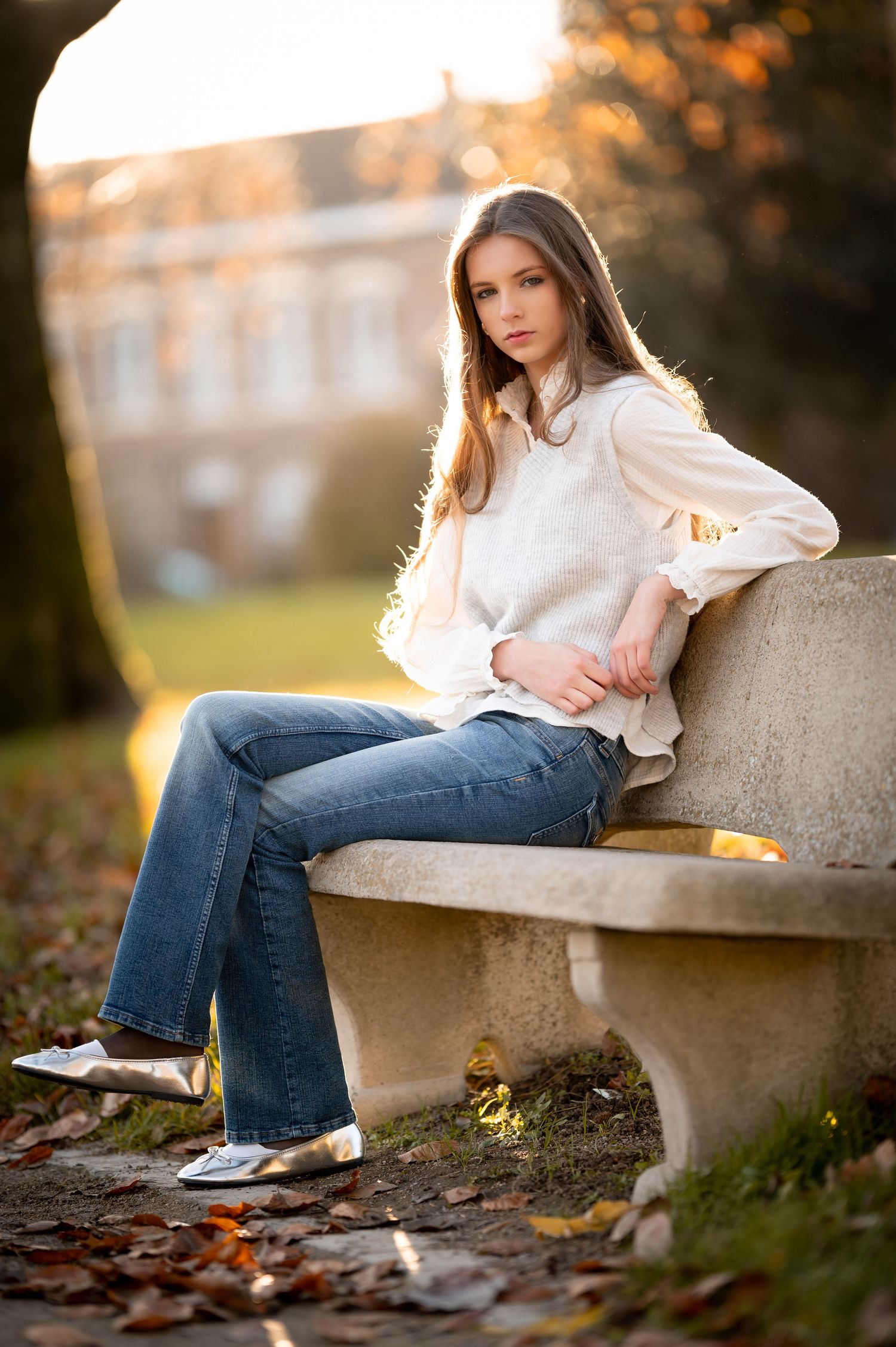 Young woman in white blouse and flare jeans sitting on a stone bench surrounded by autumn leaves.