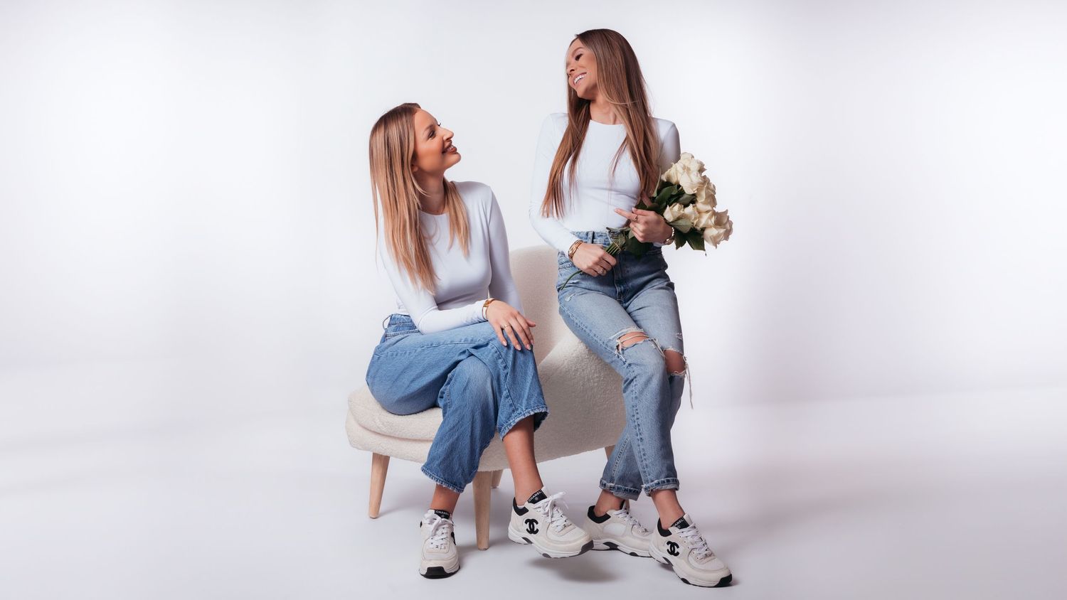 Two young women in white tops and jeans pose in a studio, one seated and one holding flowers.