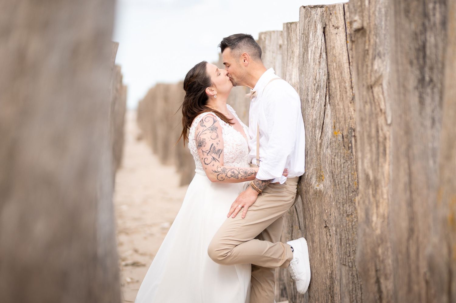 Newlyweds share a romantic kiss between rustic wooden posts on a sandy beach.