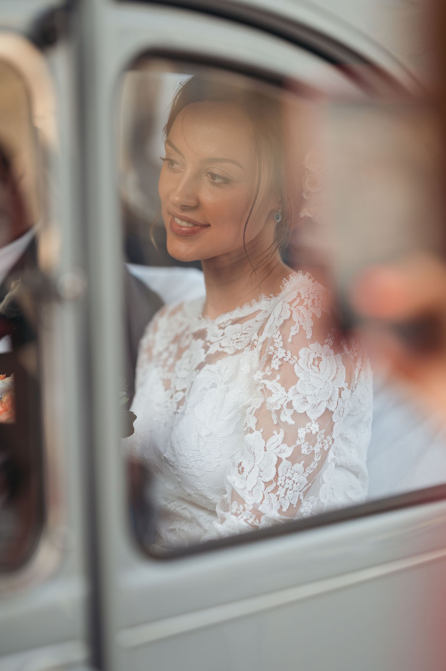 Smiling bride in elegant lace wedding dress seen through a vintage car window on her wedding day.