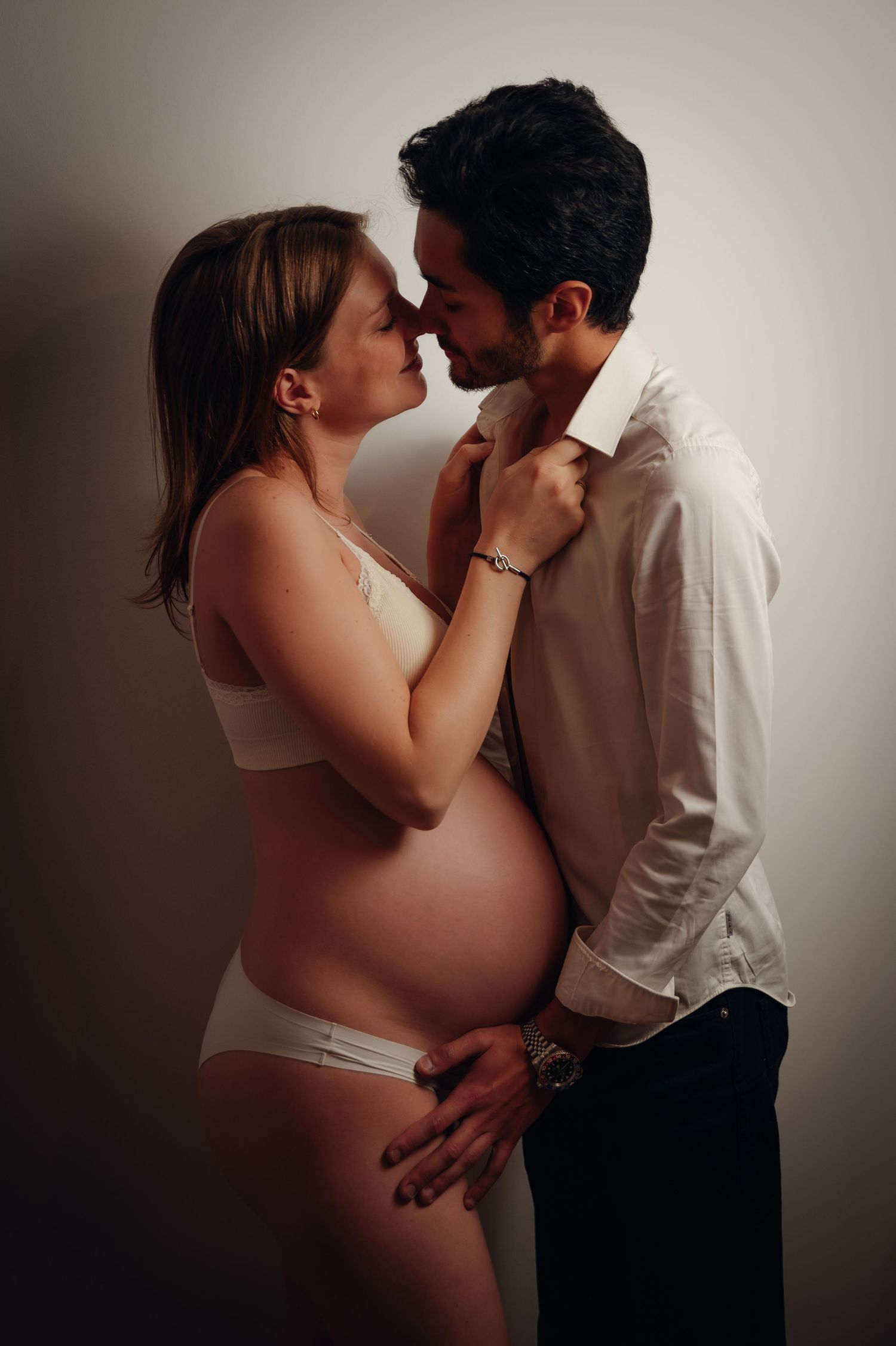 Pregnant couple sharing intimate moment in studio, woman in white lingerie showing baby bump.