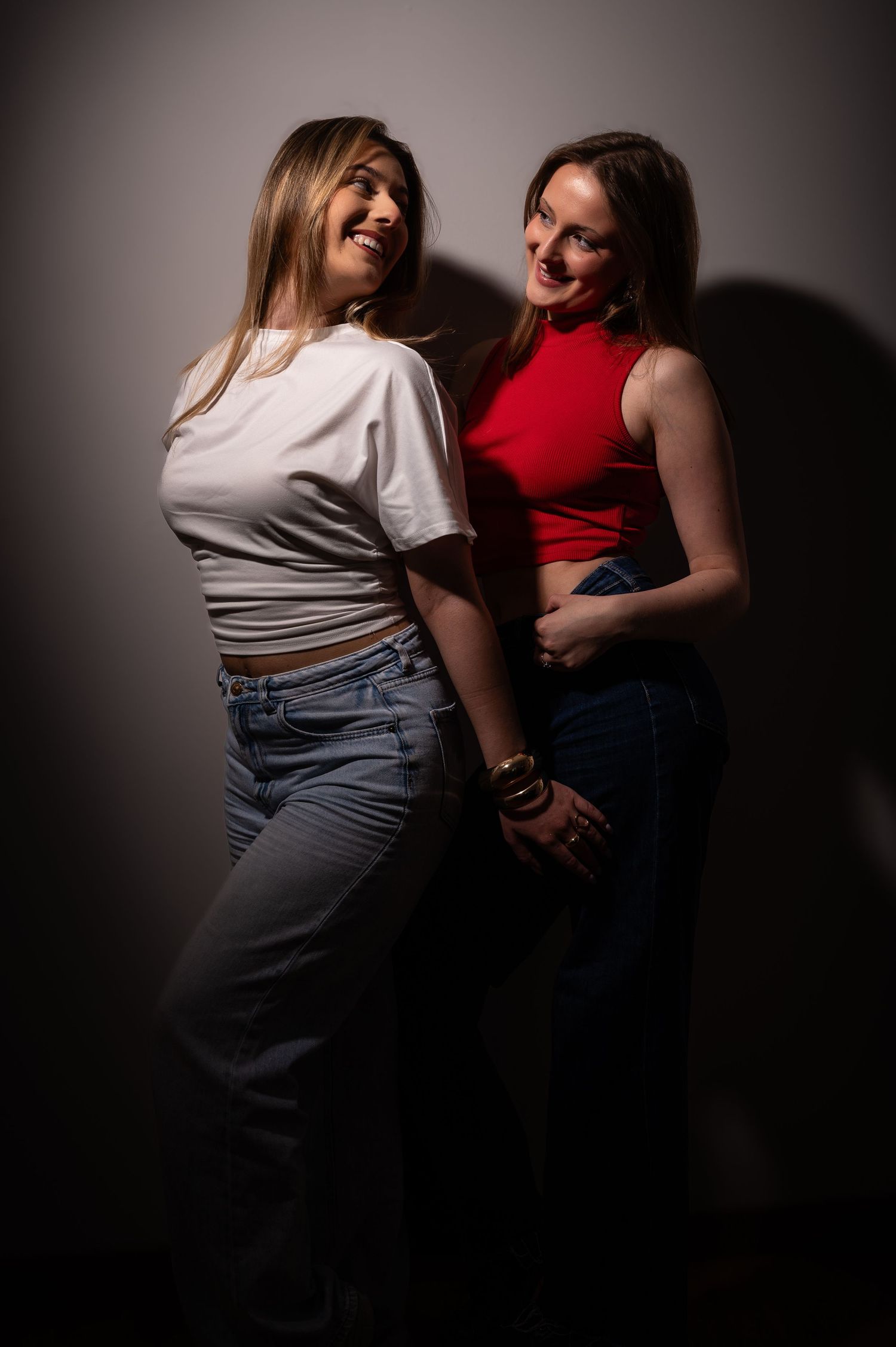 Two women smiling and posing together, one in a white crop tee and jeans, the other in a red crop top and jeans.