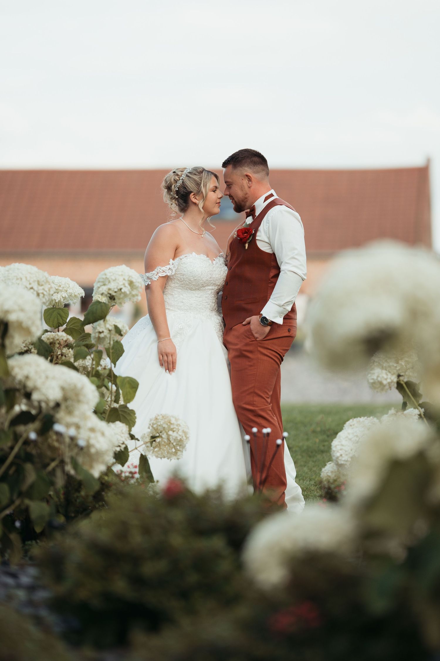 Bride in white gown and groom in rust vest share romantic moment among white hydrangeas at outdoor wedding.