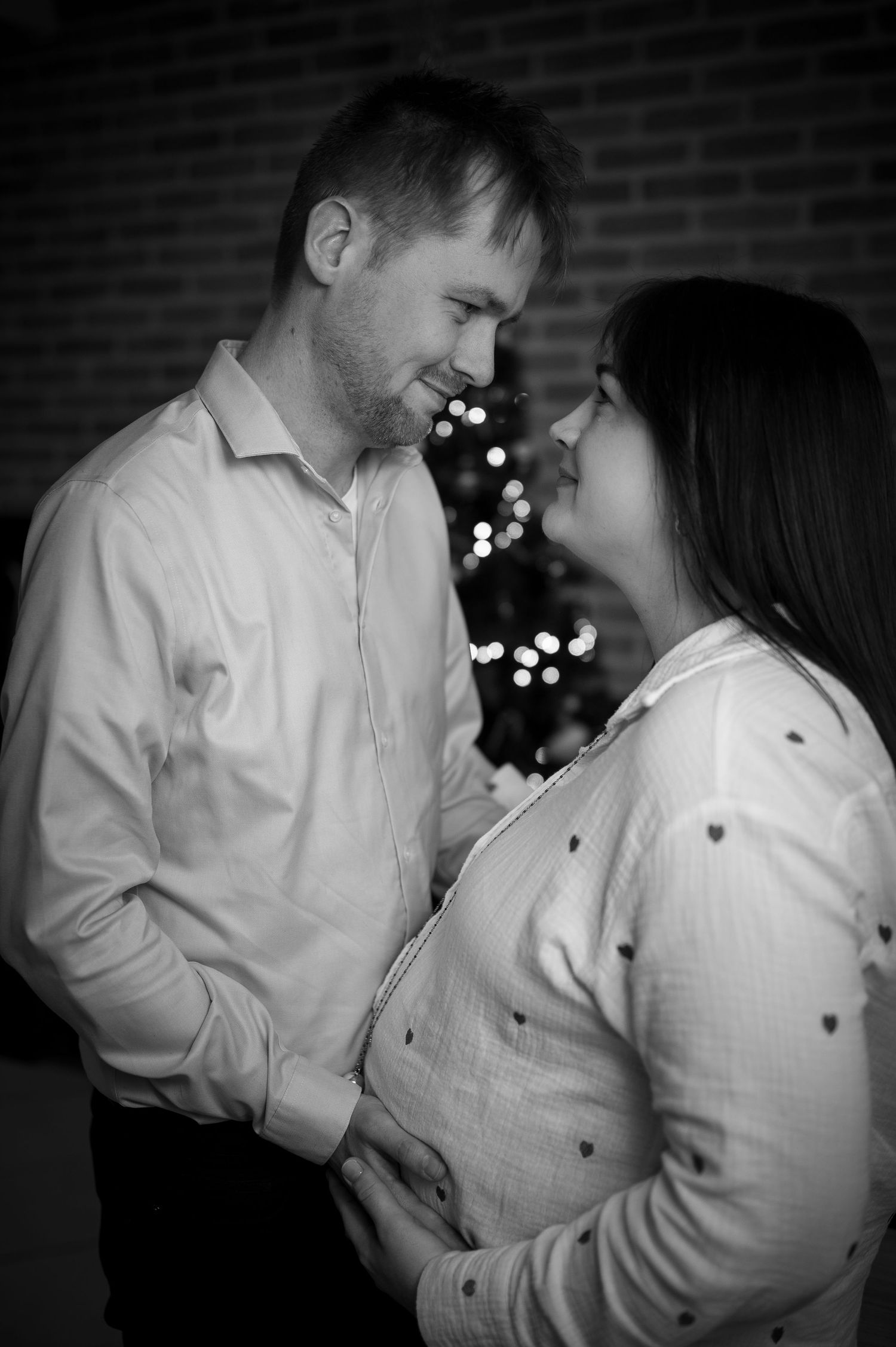 Black and white photo of a couple sharing a tender moment near a Christmas tree, with the woman visibly pregnant.