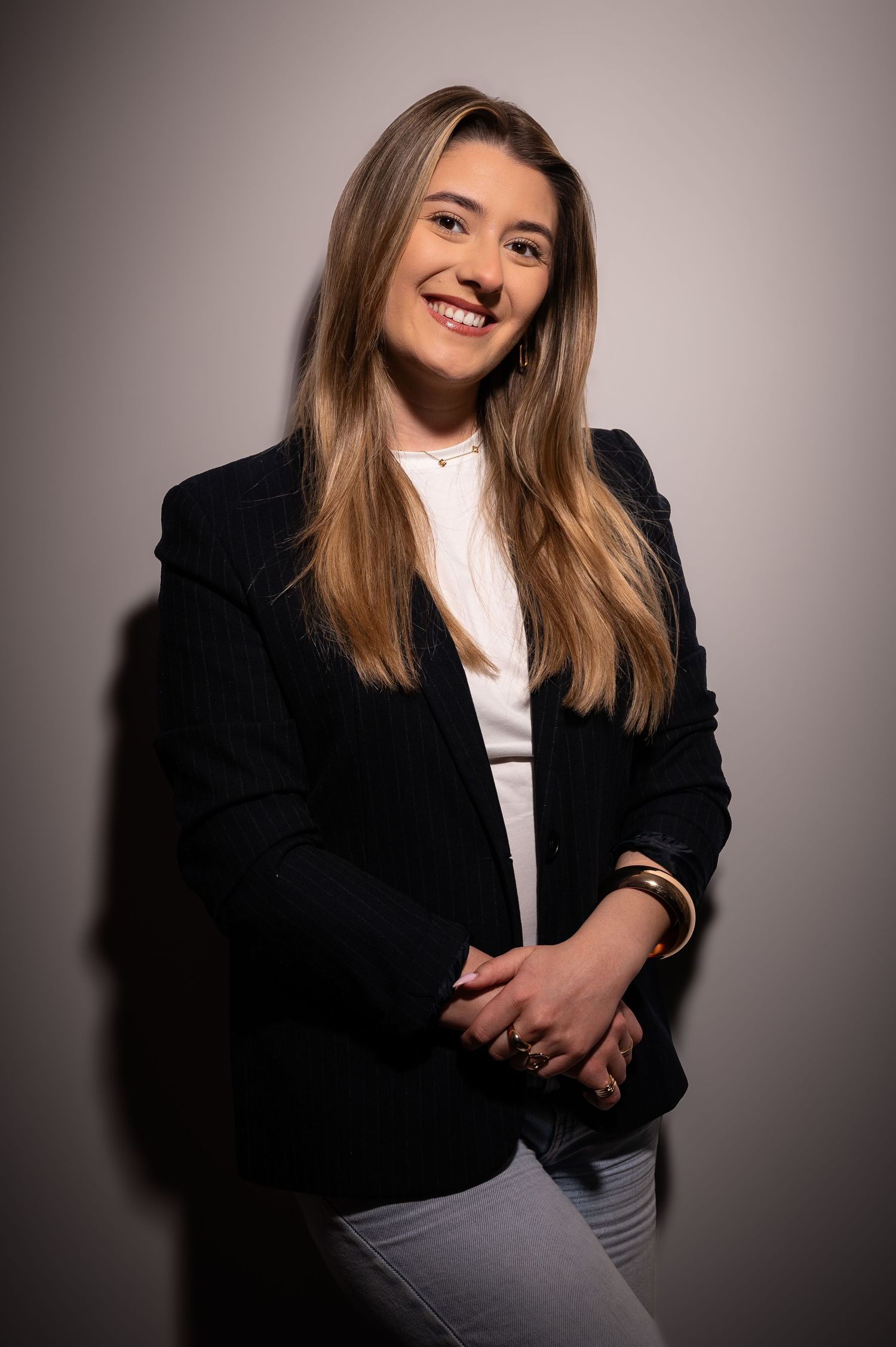 Smiling young woman with long blonde hair wearing a black blazer over white top and jeans in a professional studio portrait.