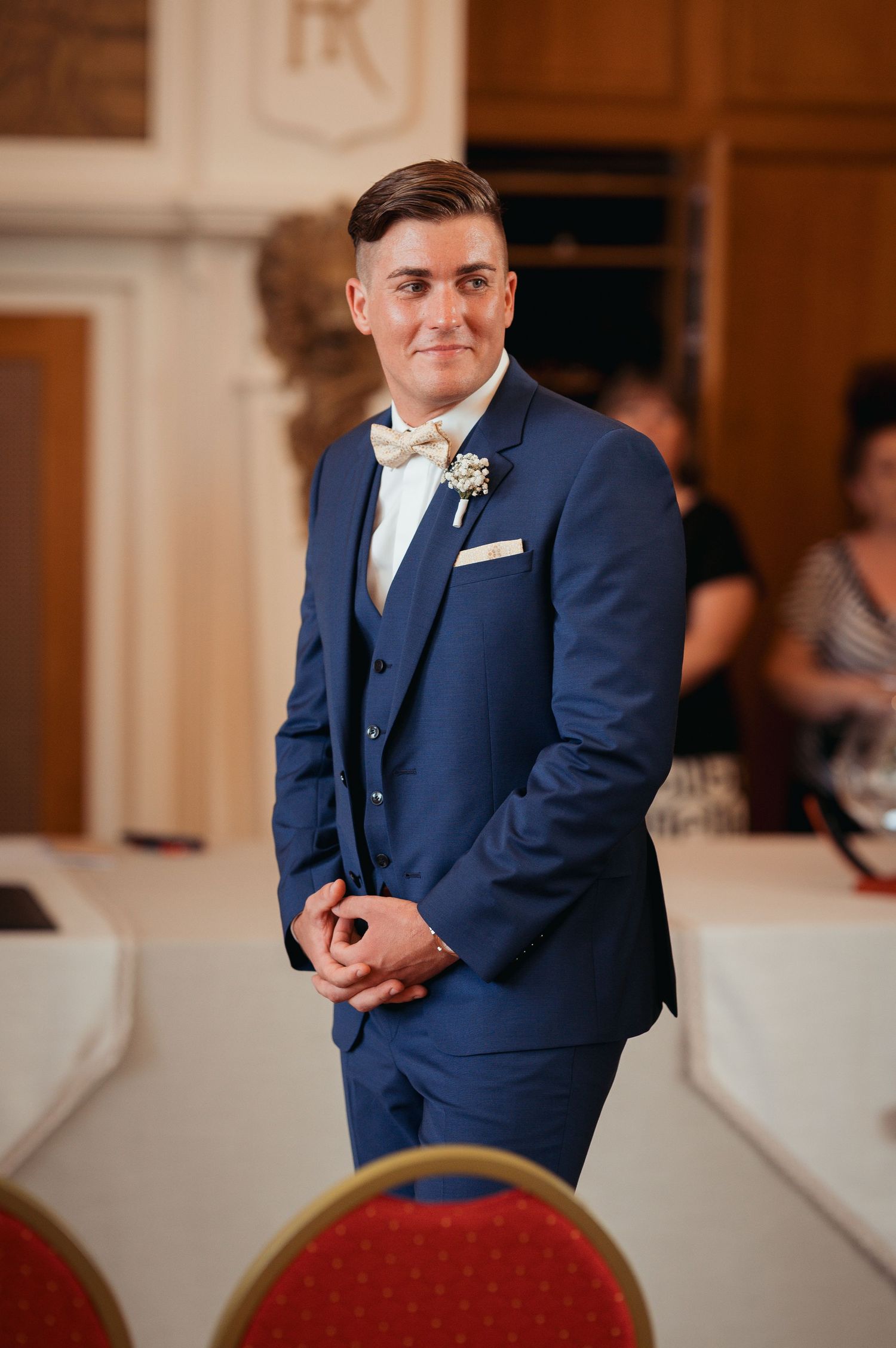 Groom in navy blue three-piece suit with bow tie and boutonniere standing at wedding venue.