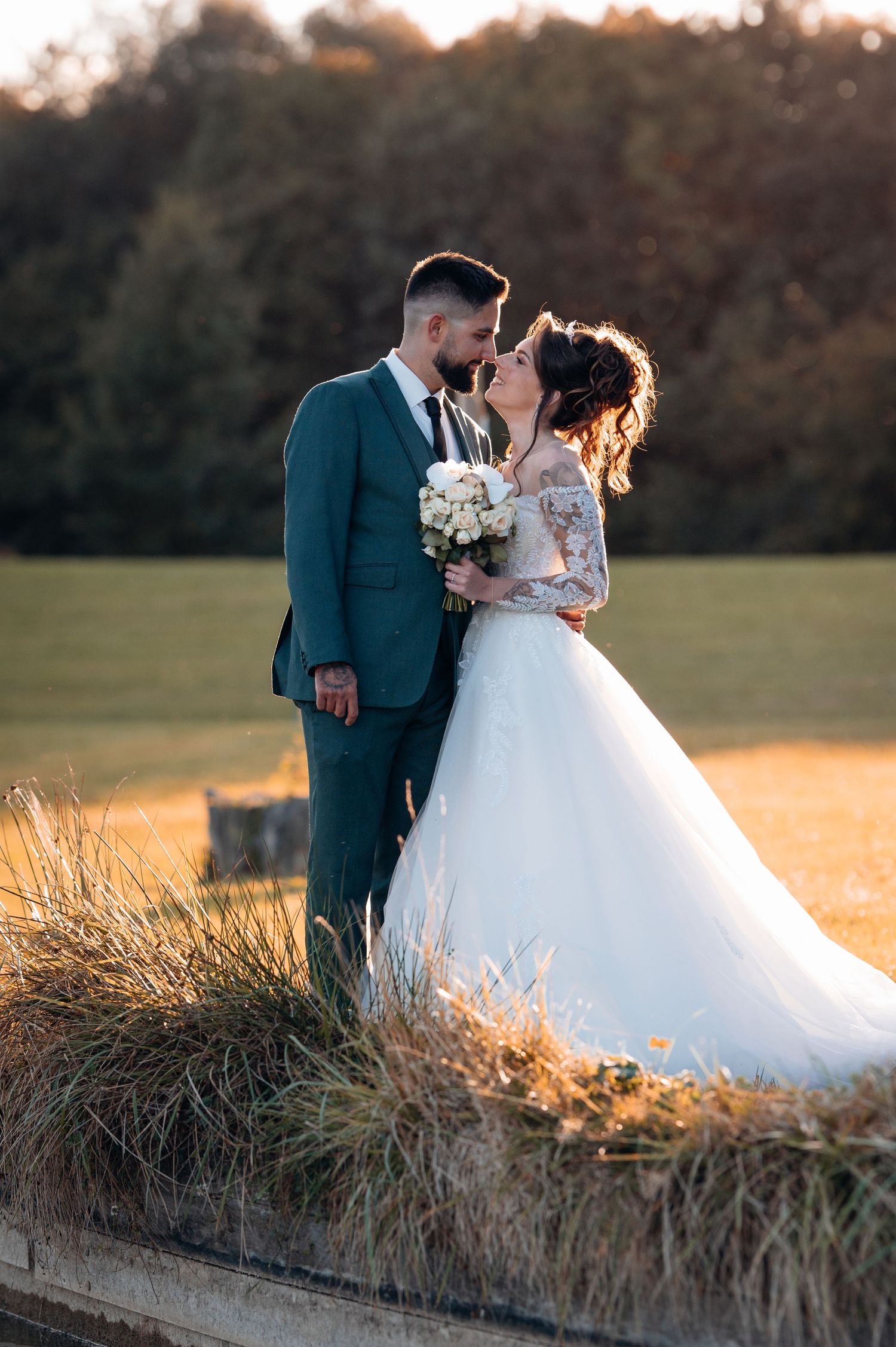 Bride and groom sharing a romantic kiss in golden hour light, with bride holding white bouquet in lush outdoor setting.