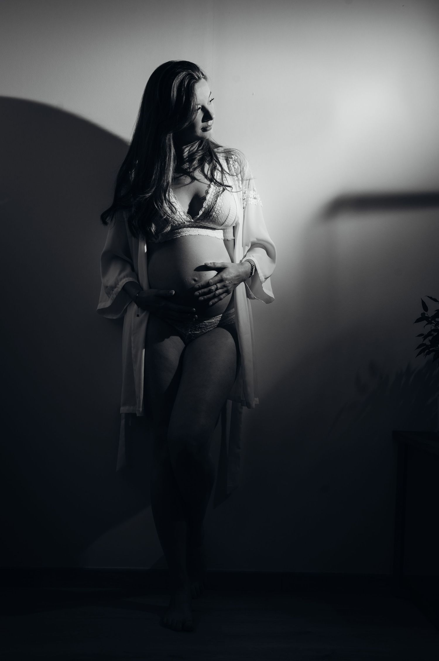 Black and white maternity photo of a woman in lingerie cradling her baby bump in dramatic studio lighting.