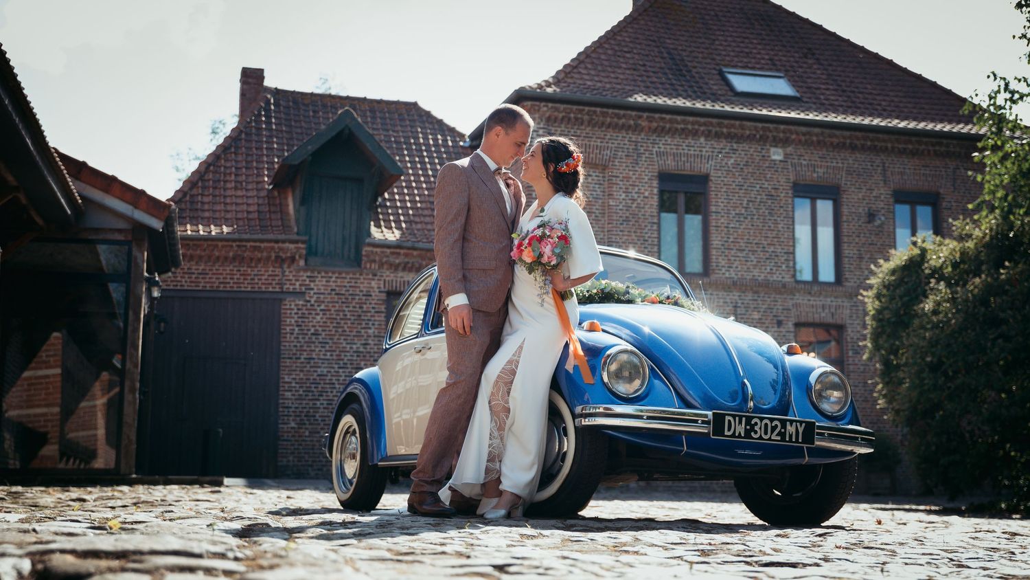 Newlyweds kiss beside a classic blue Volkswagen Beetle on cobblestone street.