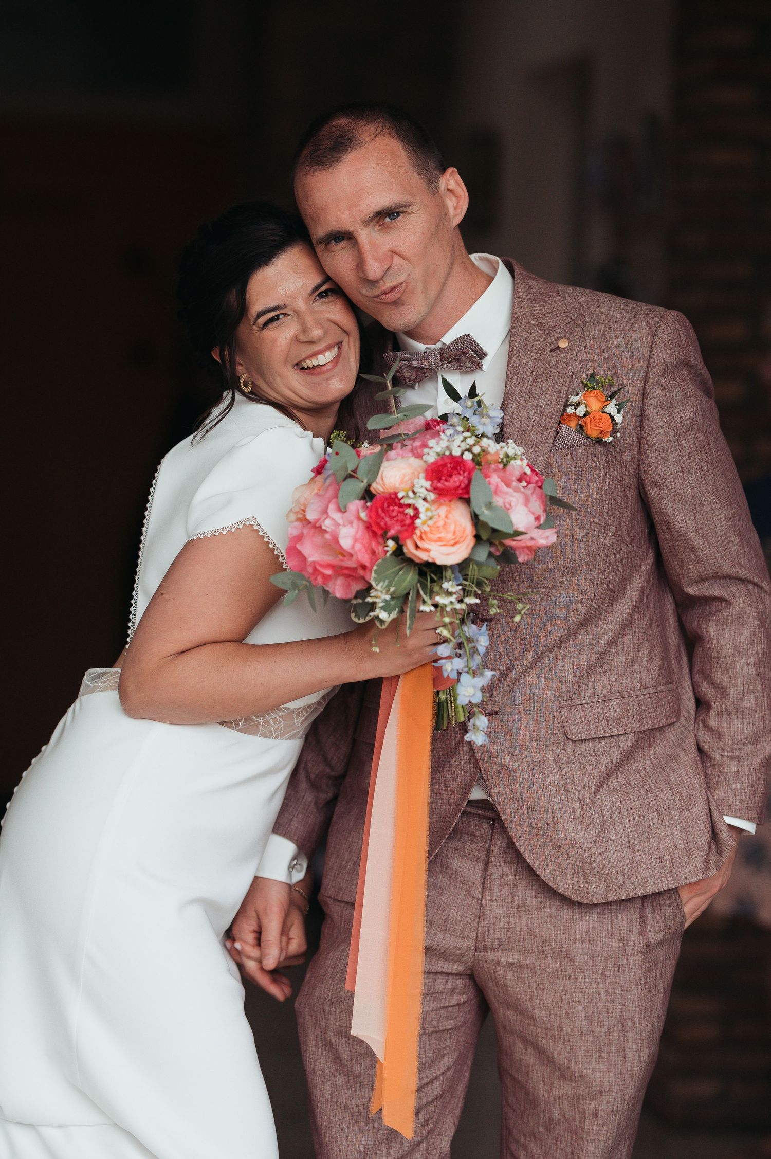 Laughing bride in white dress embraces groom in pink tweed suit holding a colorful floral bouquet.