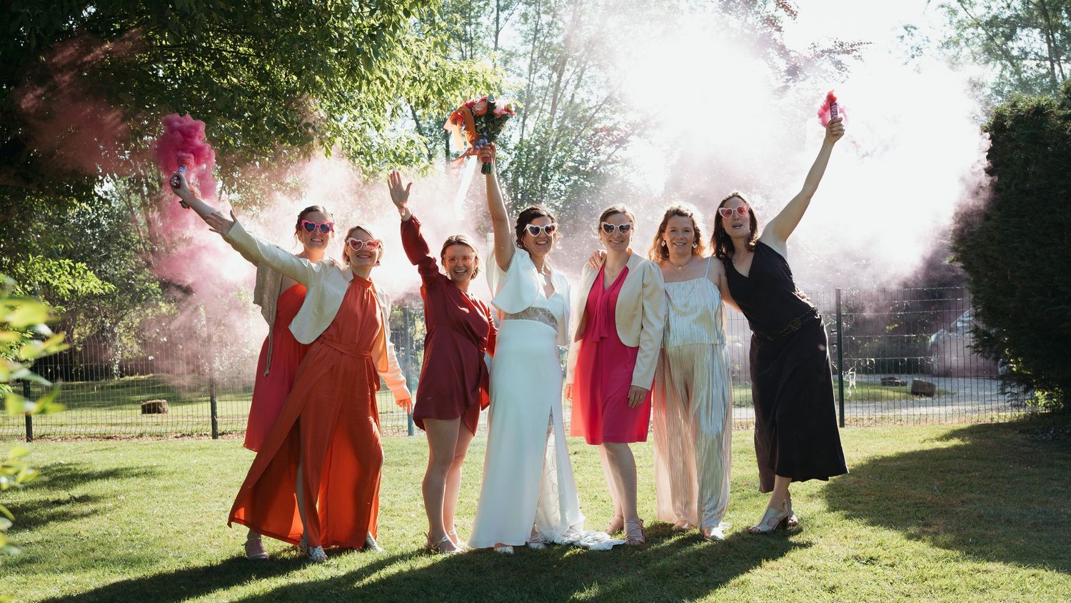 Bride and bridesmaids in colorful dresses celebrate outdoors with pink and purple smoke bombs on a sunny day.