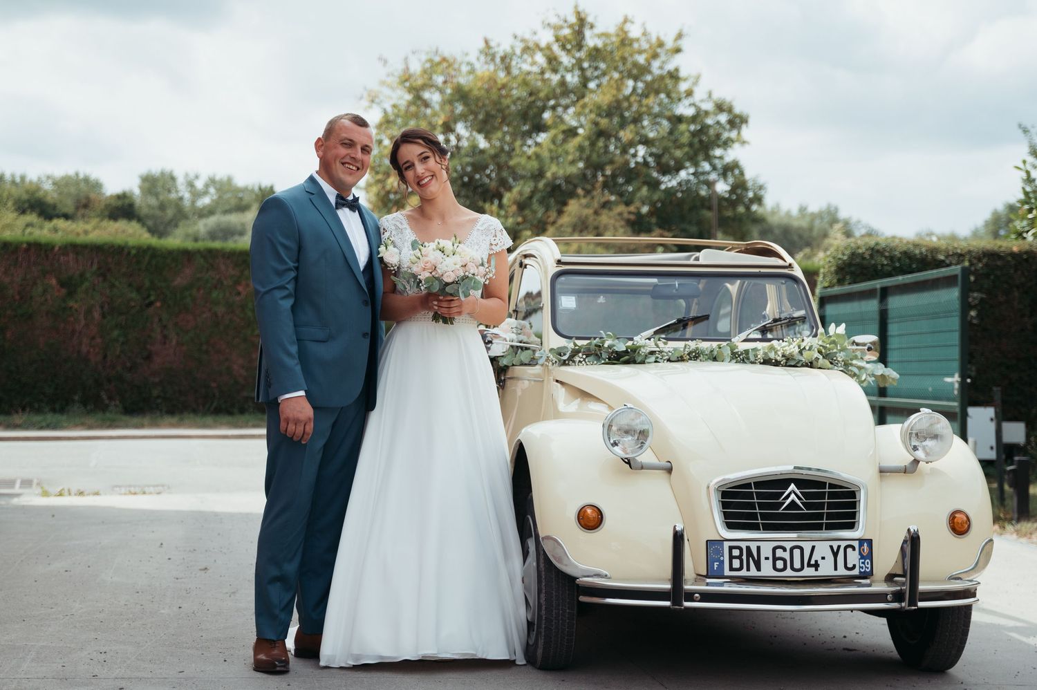 Bride and groom posing beside a vintage cream Citroen 2CV decorated with flowers at a French wedding.