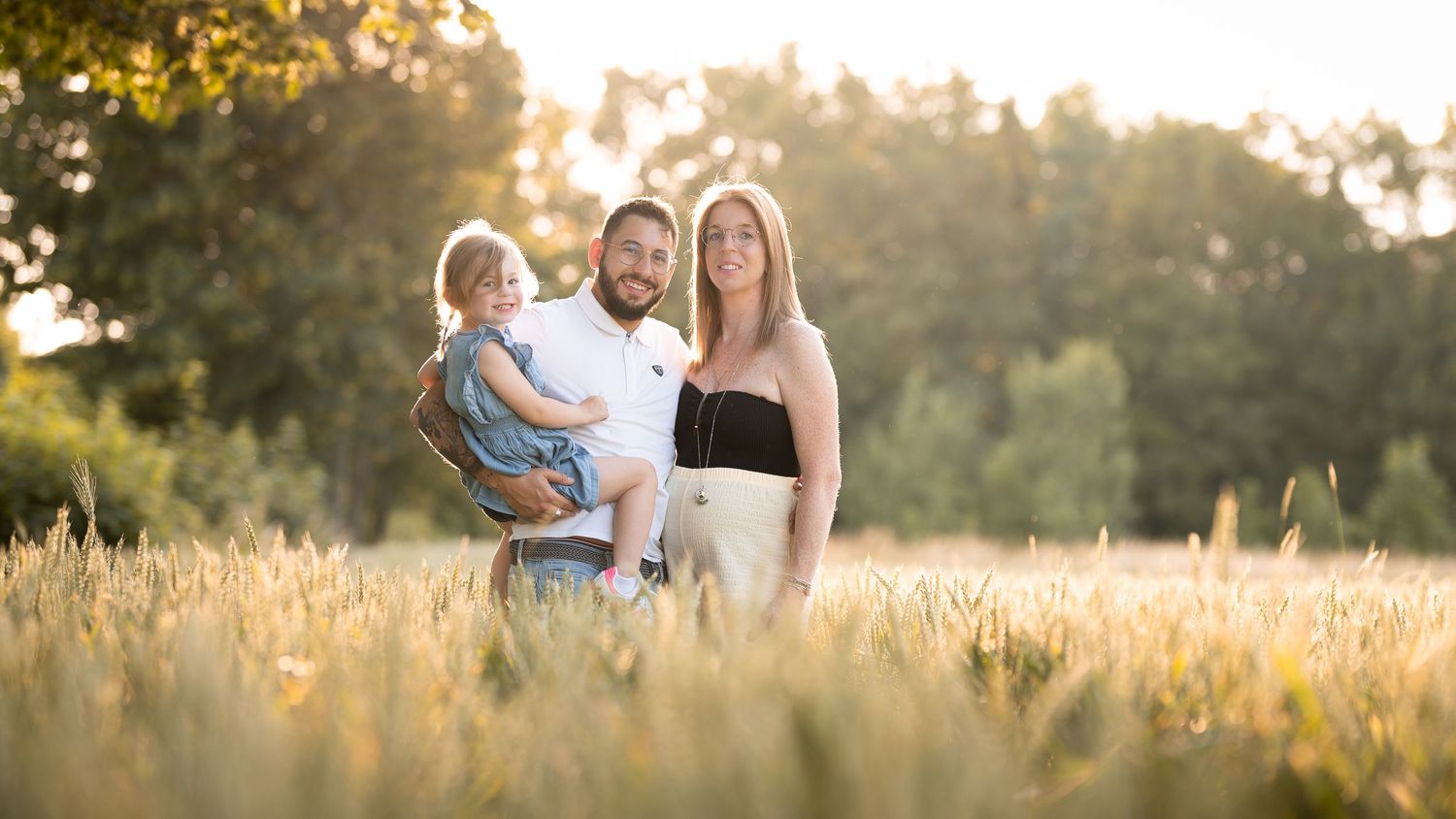 Family of three posing in golden wheat field at sunset, parents holding toddler with trees in background.