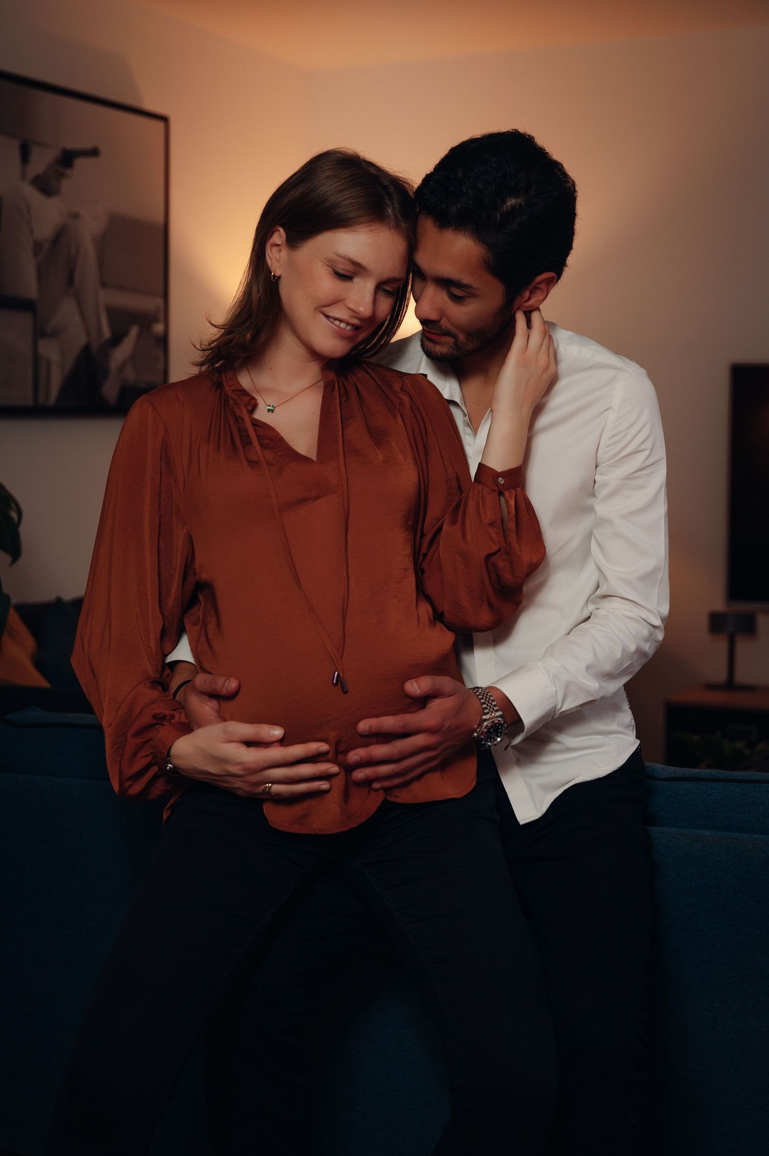 Couple sharing tender moment, man embracing pregnant woman from behind, both smiling in warm dimly lit room.