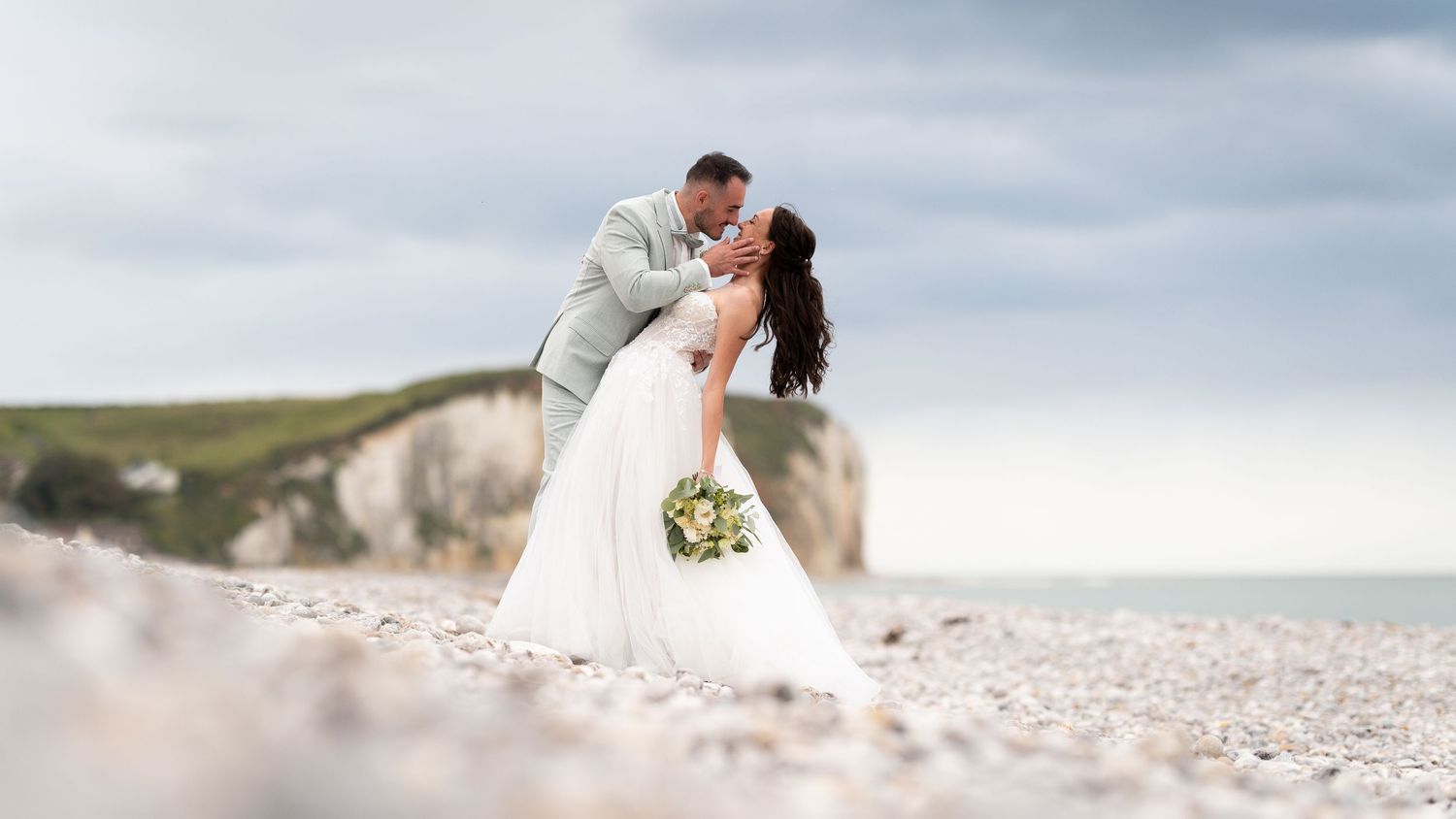 Bride and groom share a romantic dip kiss on a pebble beach with white cliffs in the background.