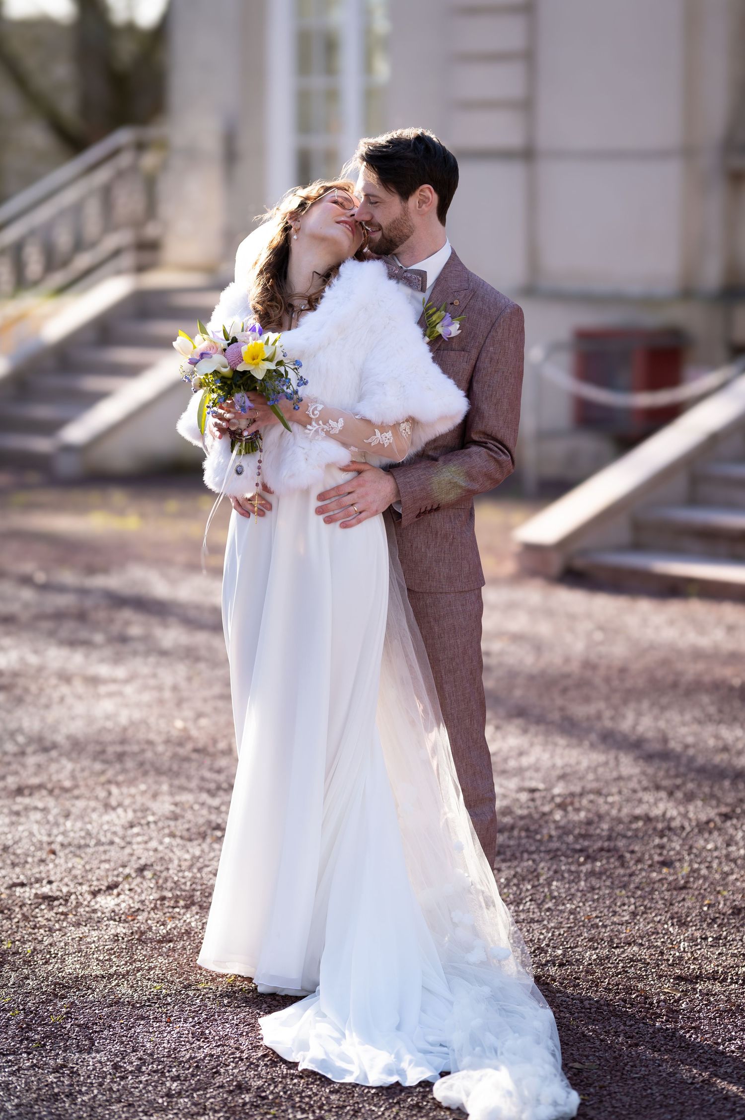 Wedding couple embrace outdoors, bride in white gown with fur wrap holding spring bouquet, groom in brown tweed suit.