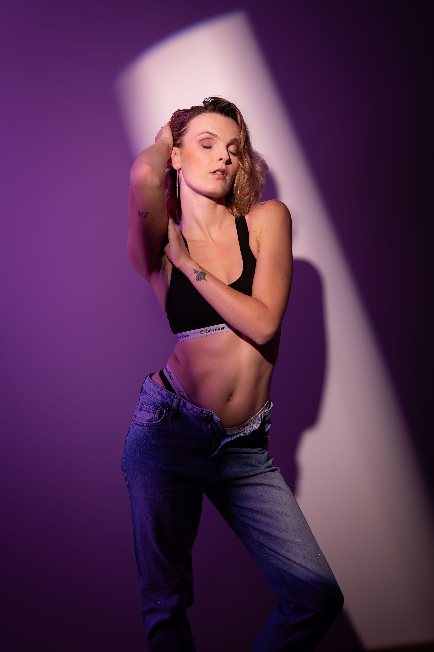 Athletic woman posing in black sports bra and low-rise jeans against a purple background with dramatic studio lighting.