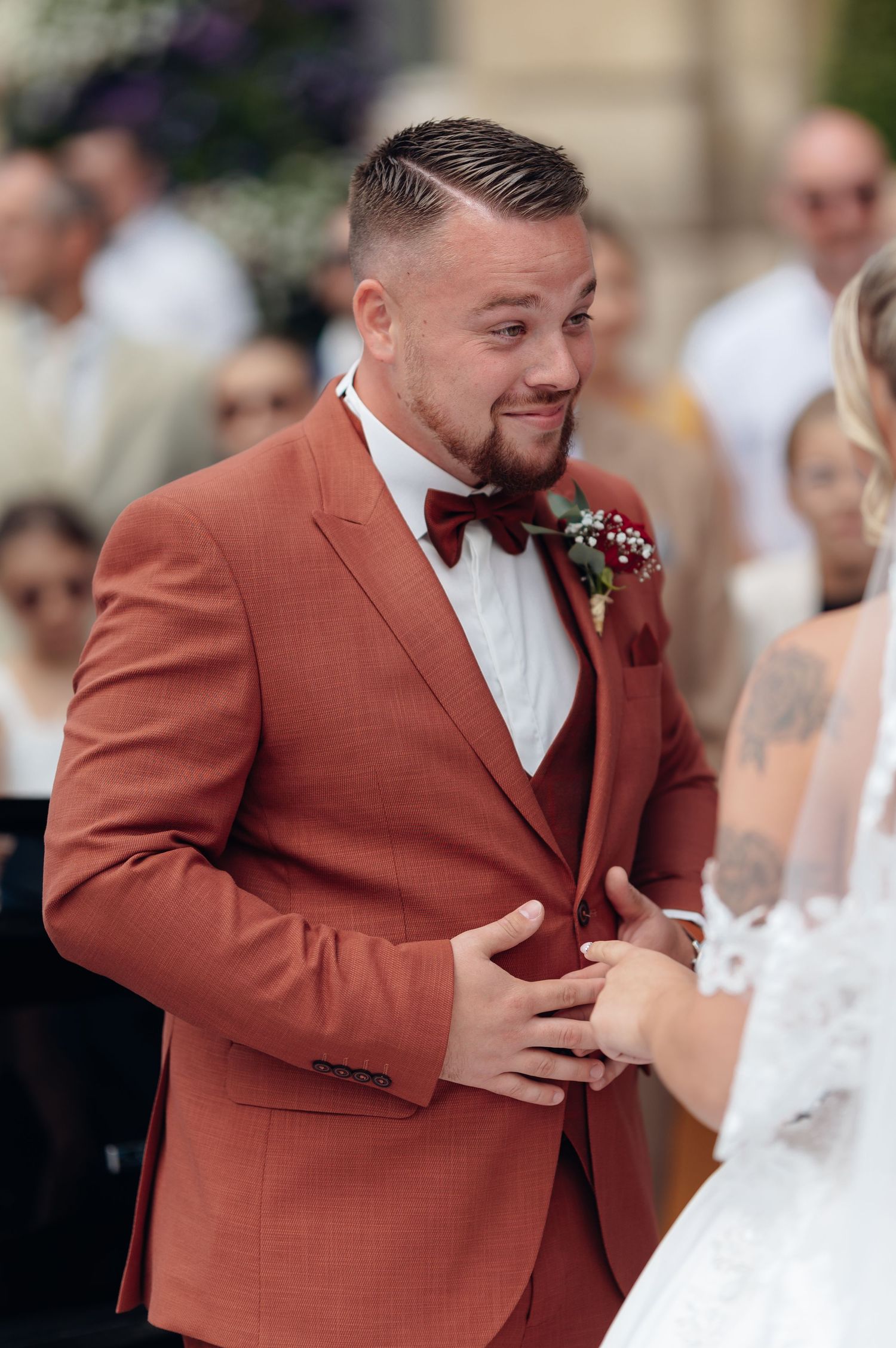 Groom in rust-colored three-piece suit with bow tie smiling emotionally at bride during outdoor wedding ceremony.