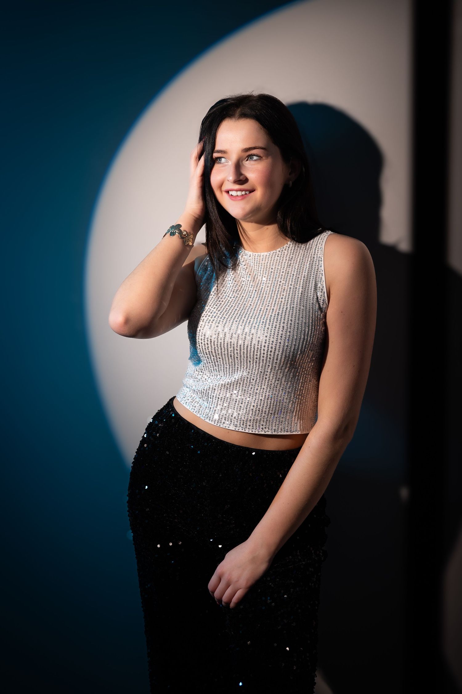 Smiling young woman in silver crop top and black sequin skirt posing against teal wall with spotlight.