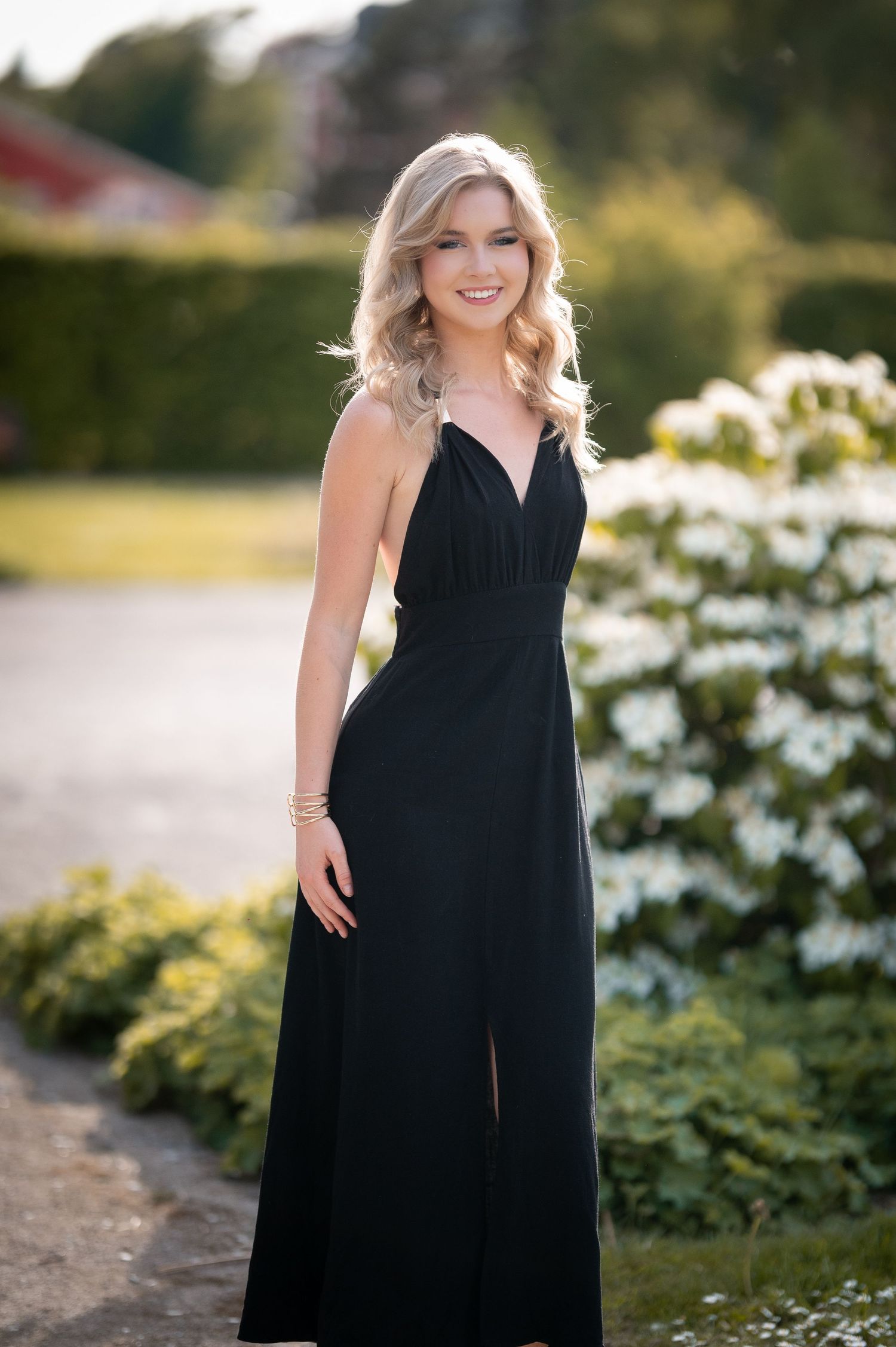 Young blonde woman smiling in elegant black evening gown outdoors, surrounded by lush greenery and white flowers.