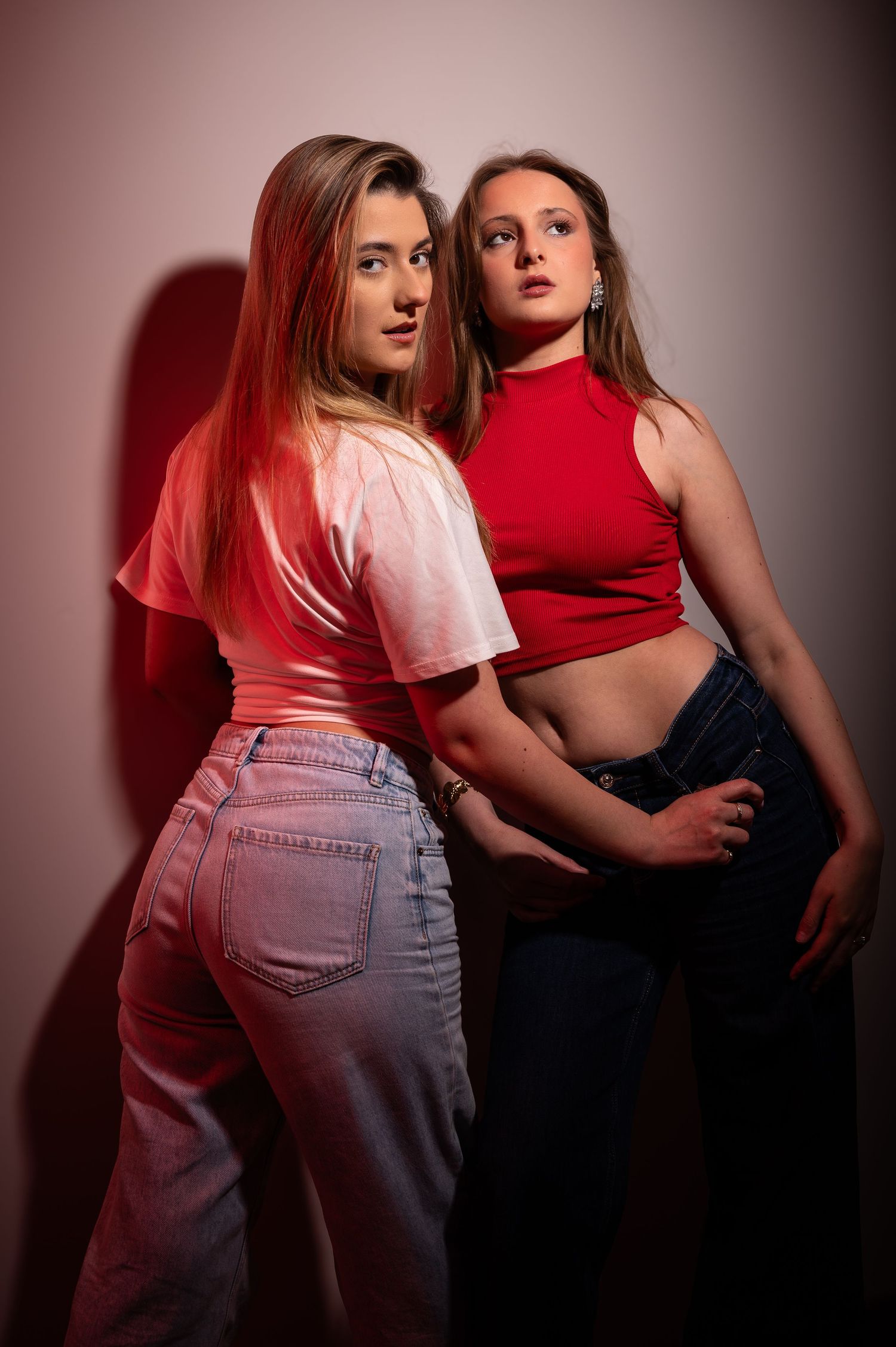 Two young women in red tops and jeans pose together against a pink wall in a stylish fashion portrait.