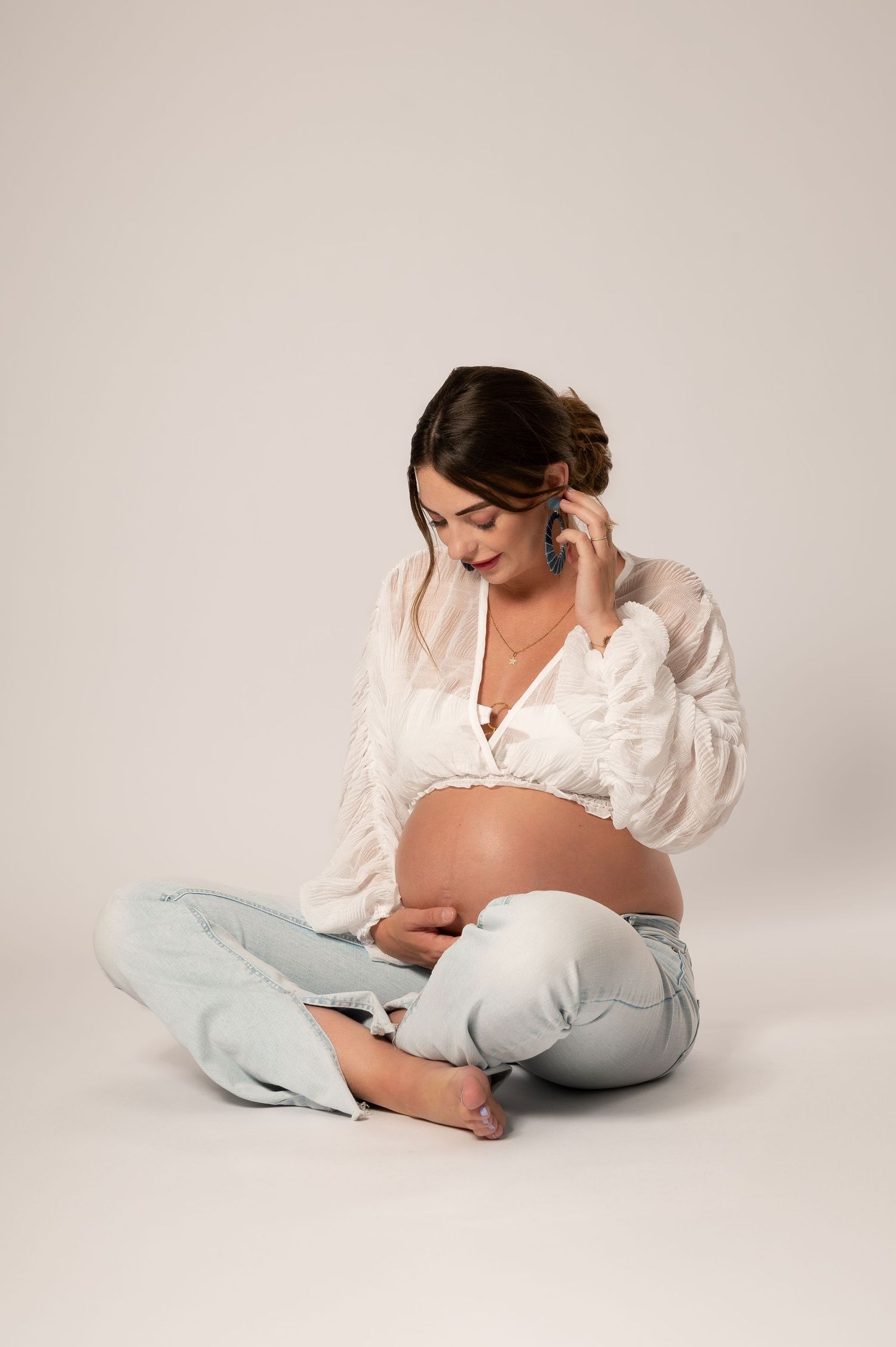 Pregnant woman sitting cross-legged on floor, talking on phone, wearing white lace top and light blue pants.