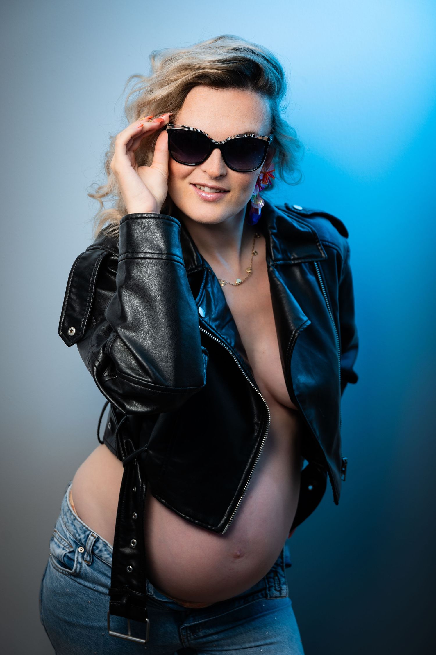 Pregnant woman in leather jacket and sunglasses posing confidently against a blue studio background.