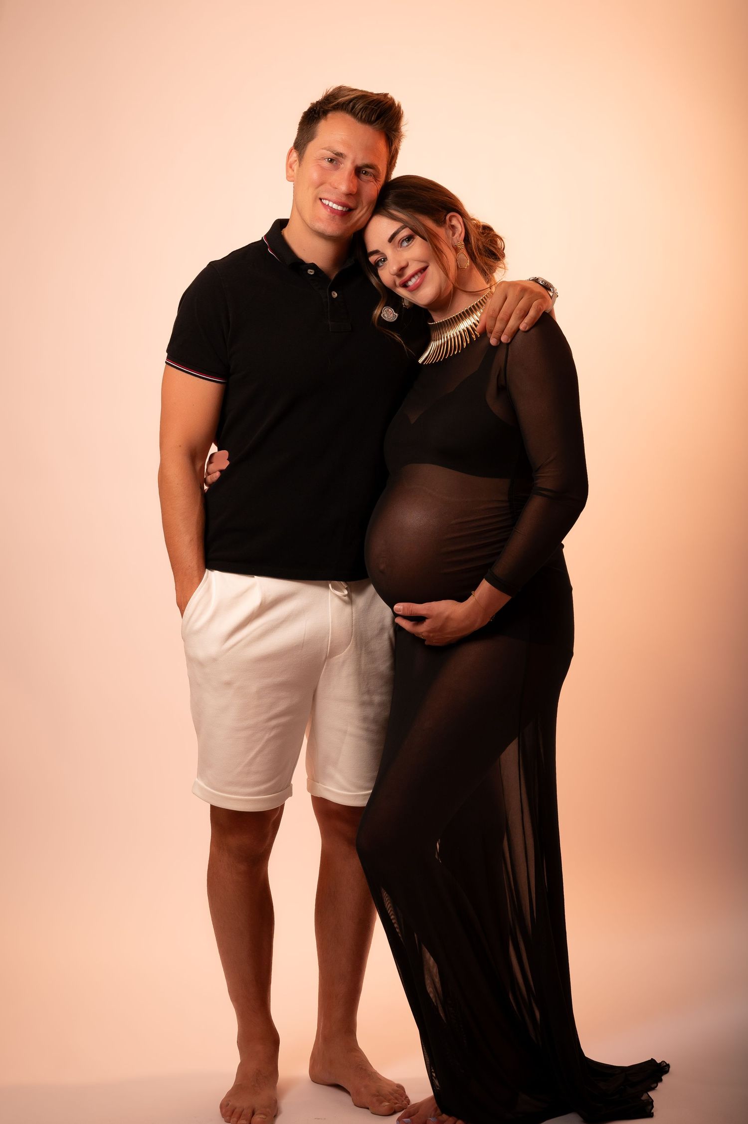 A smiling couple poses together, the woman in a black sheer maternity gown cradling her baby bump, the man in a black polo and white shorts.