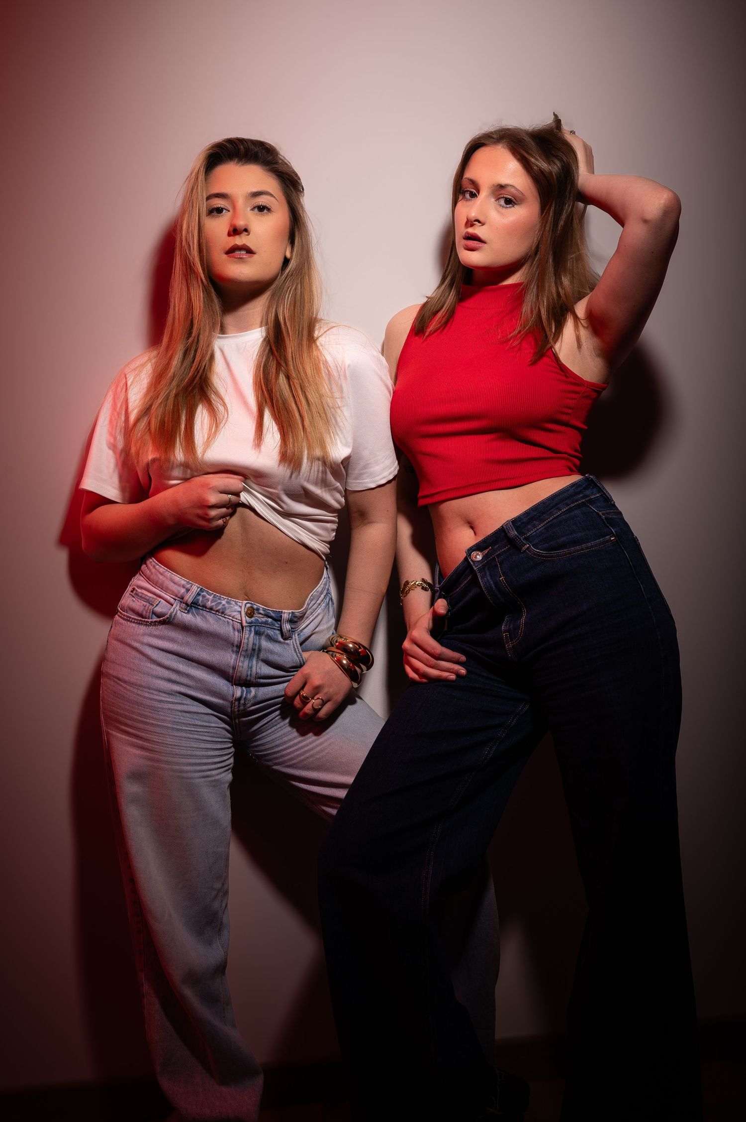 Two young women posing in casual crop tops and jeans under red and white studio lighting.