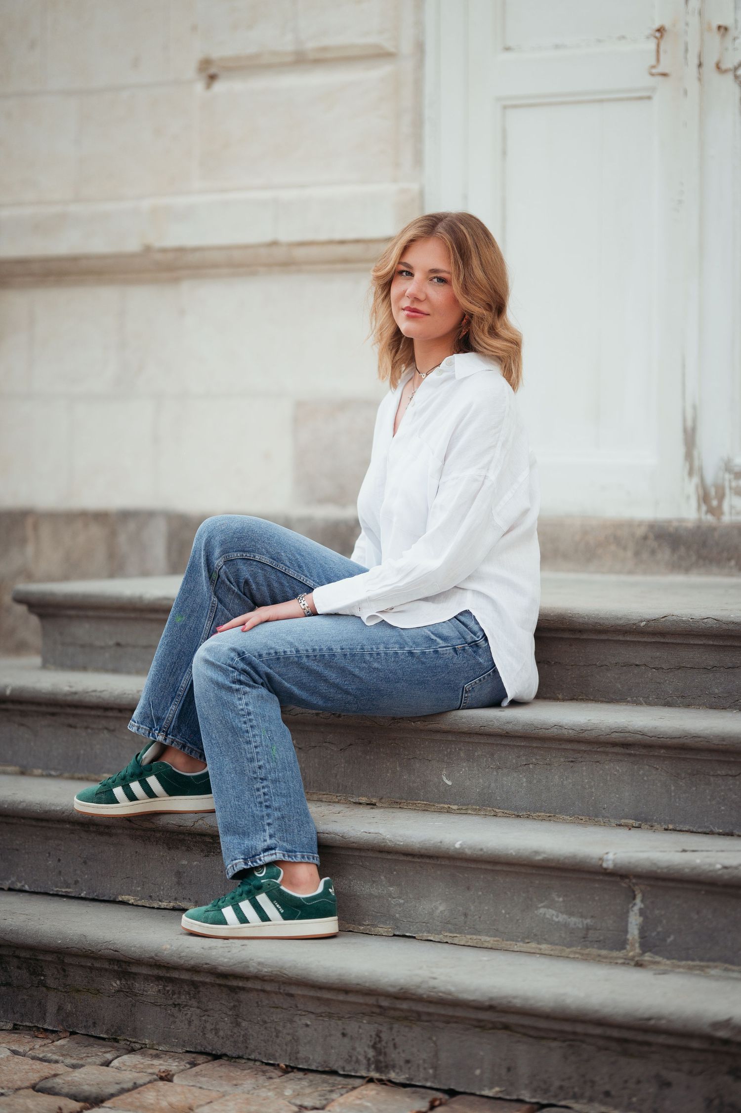 Woman in white shirt, blue jeans, and green Adidas sneakers sitting on stone steps outdoors.