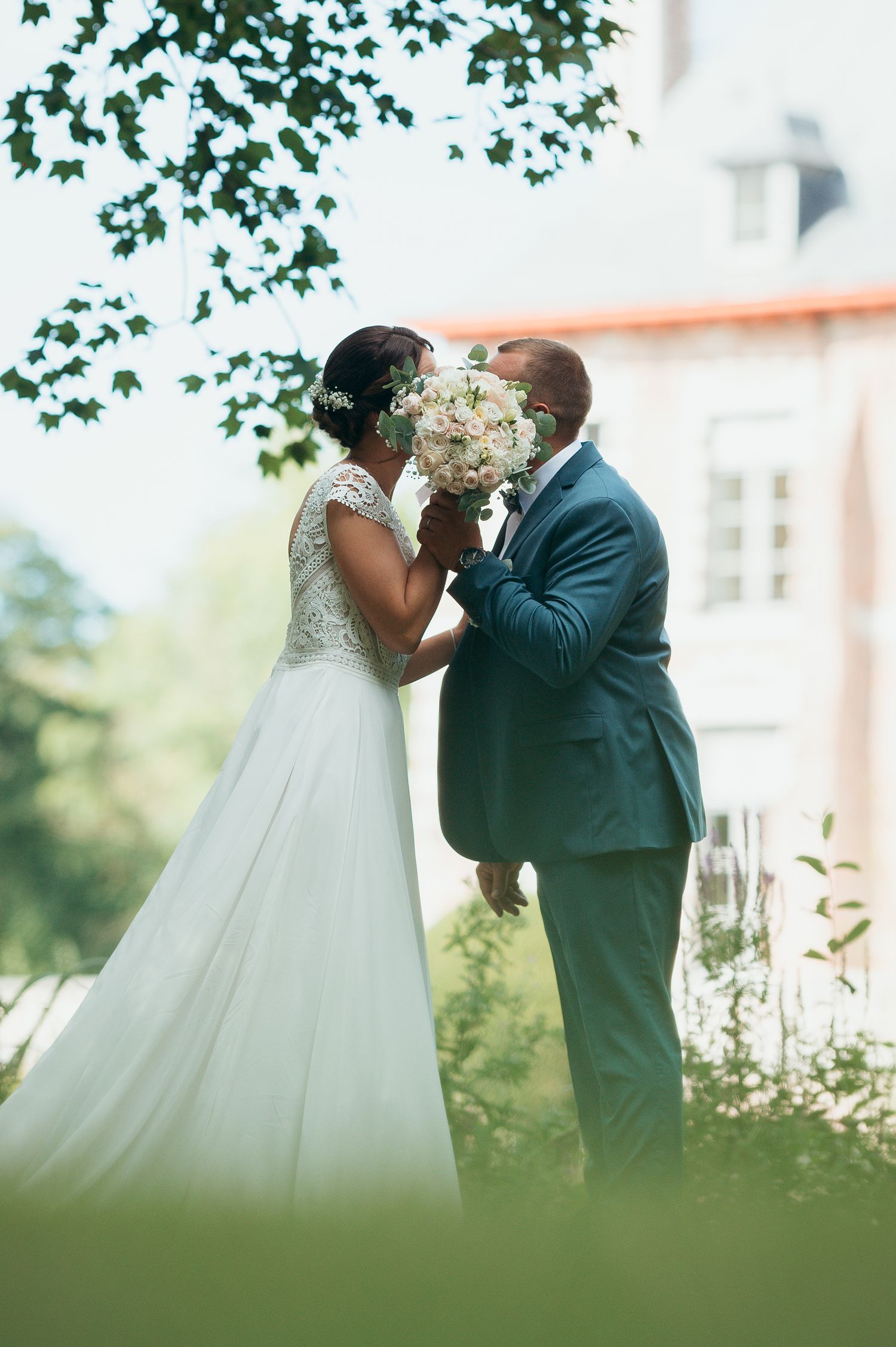 Bride and groom share a romantic kiss behind a white bouquet outdoors, with a grand building and lush greenery in the background.