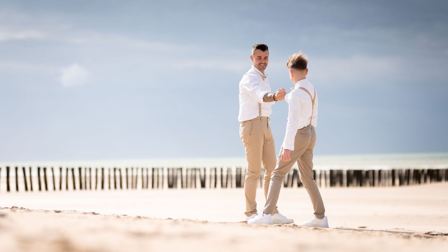 Deux hommes père et fils en chemises blanches et pantalons beiges se tiennent la main sur une plage de sable sous un ciel bleu.