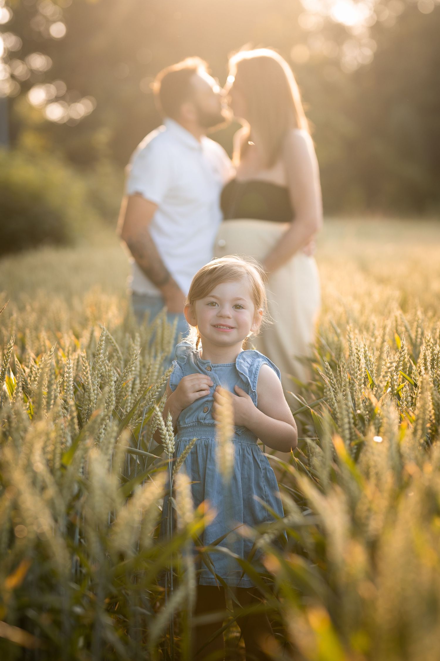 Smiling toddler girl in blue dress stands in golden wheat field with kissing parents.