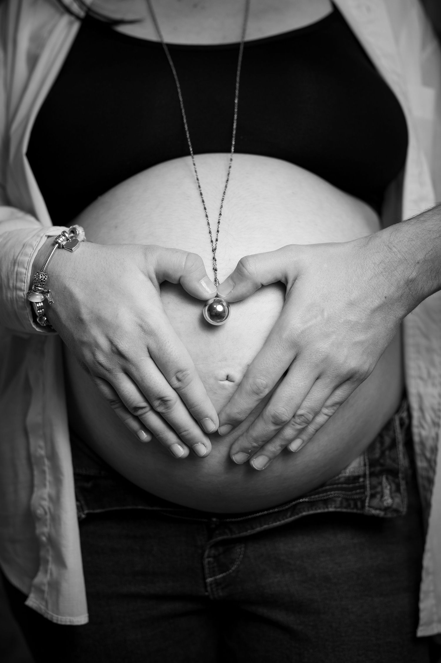 Black and white maternity photo of pregnant woman with hands forming heart shape over baby bump, wearing necklace and jeans.