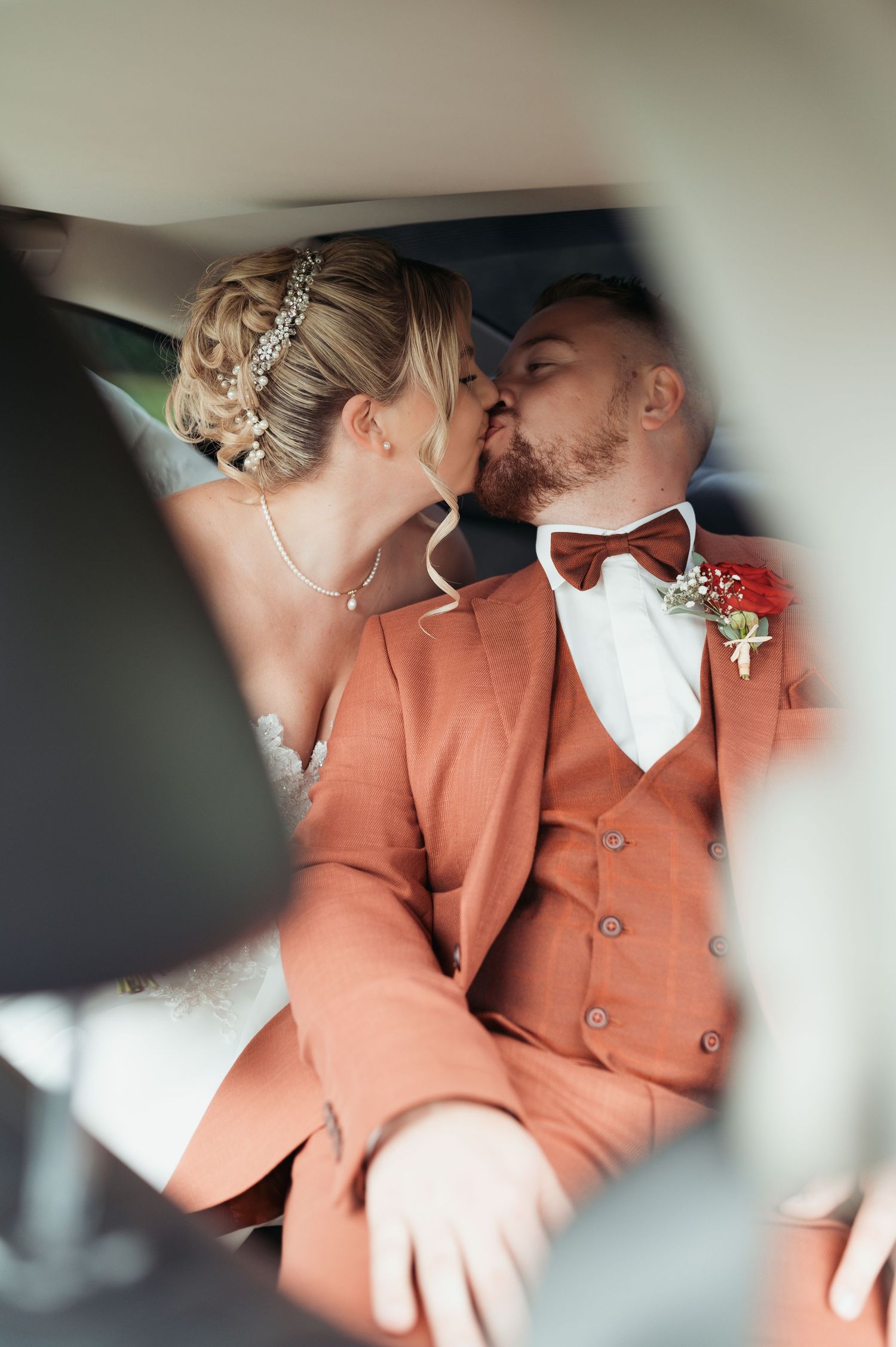 Bride and groom sharing a romantic kiss in a car, groom wearing a terracotta suit with bow tie and boutonniere.