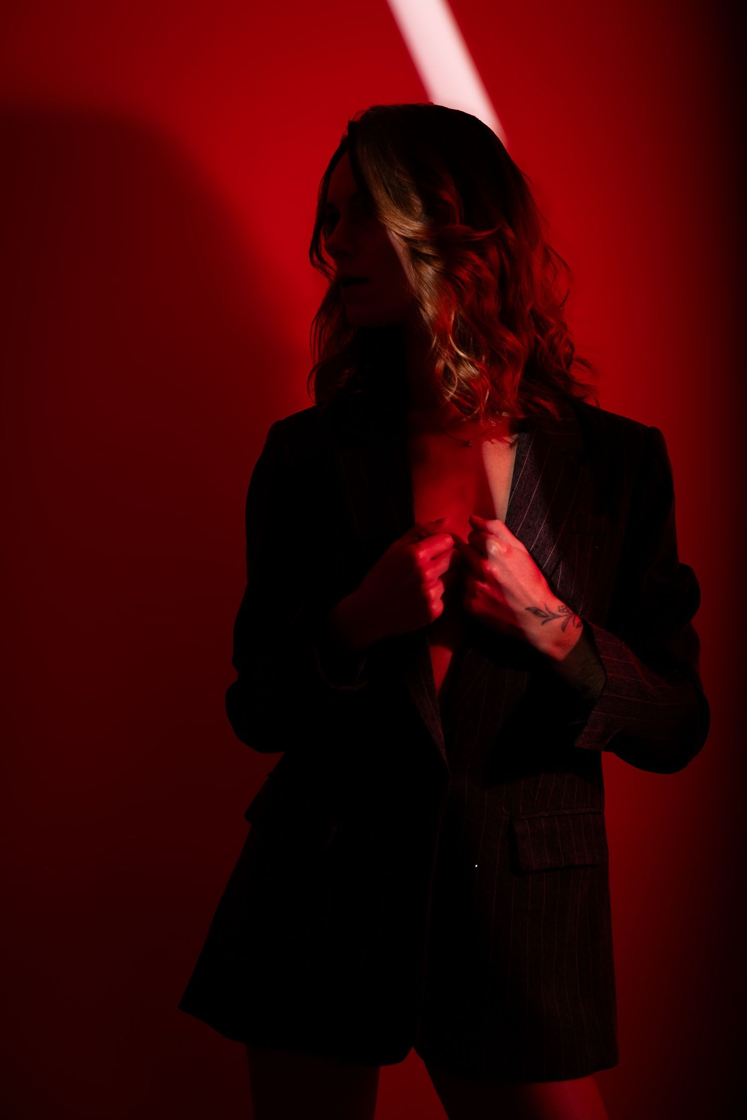 Woman in dark blazer posing against a red background with dramatic lighting highlighting tattoos.