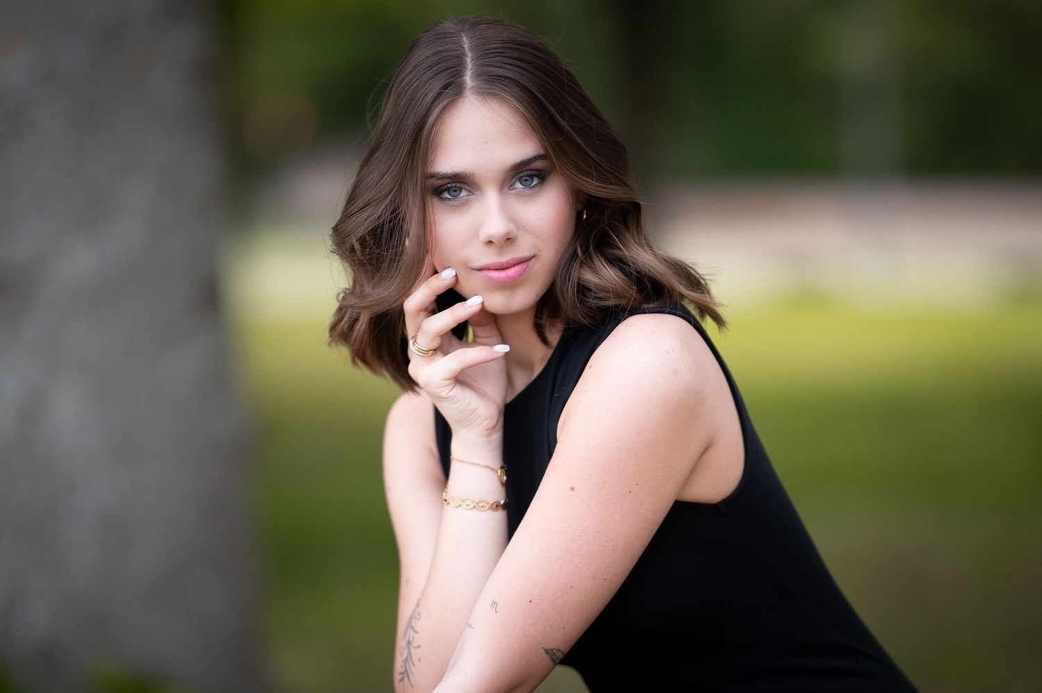 Young woman in black dress poses outdoors in park, looking over shoulder with hand near face.