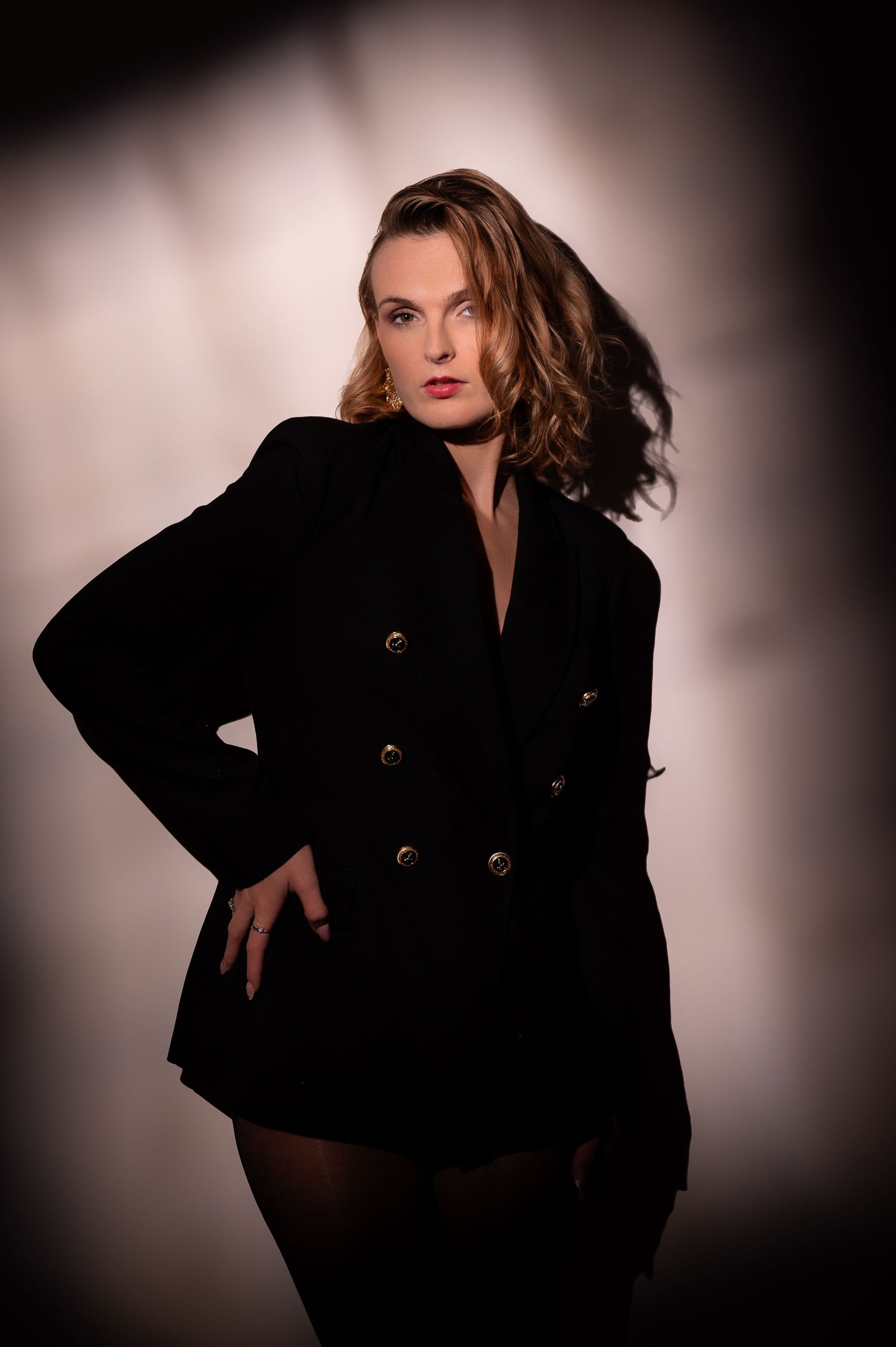 Stylish woman in oversized black double-breasted blazer dress posing confidently in a dramatic studio lighting setup.