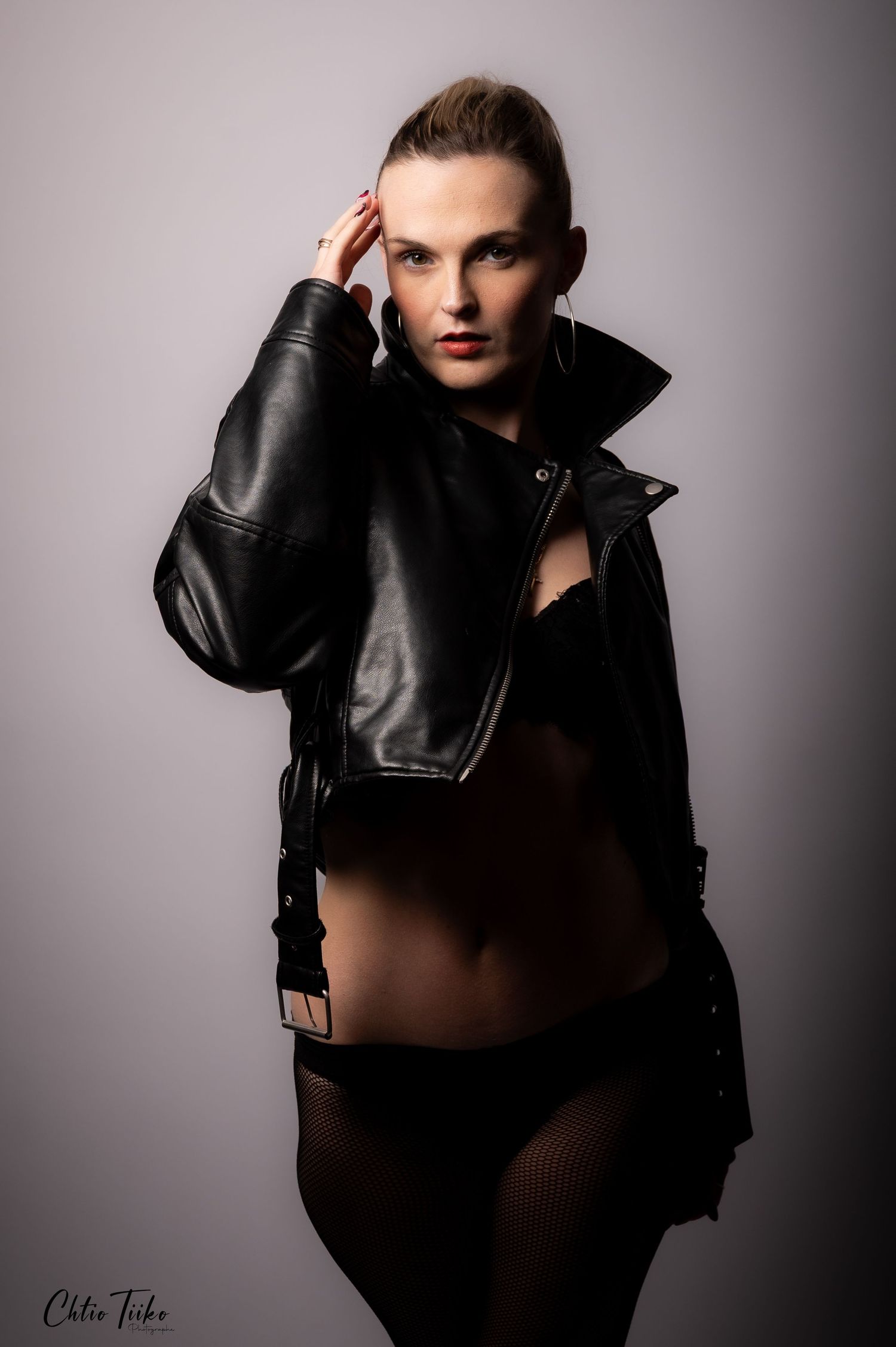 A woman poses confidently in a black leather jacket and lingerie against a neutral studio background.