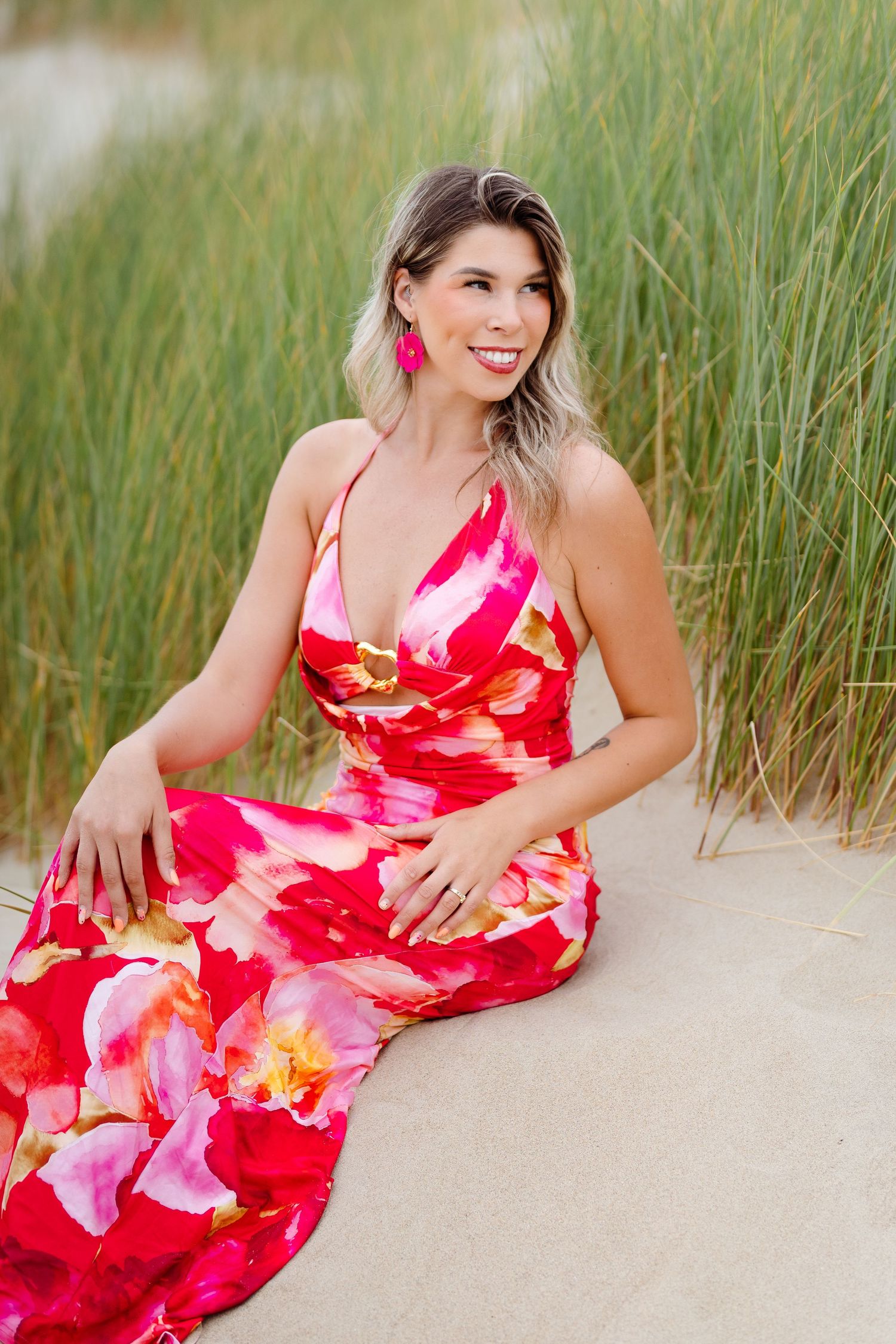 Woman in red floral maxi dress sitting on sandy beach with tall grass, smiling, wearing pink earrings.